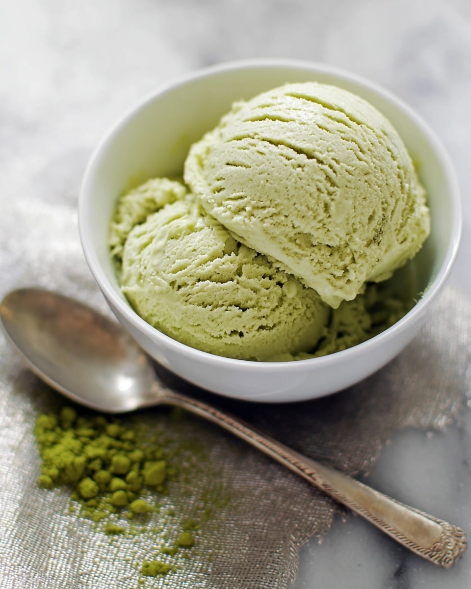 A close-up view of a white bowl filled with two scoops of pale green ice cream. The ice cream has a smooth and slightly crumbly texture with visible lines from scooping on top. The bowl sits on a white marbled surface with some loose green powder scattered to the left. A silver spoon with a rounded handle lies next to the bowl on a softly textured metallic cloth, creating a simple and fresh presentation. photo taken with an iphone --ar 4:5 --v 7