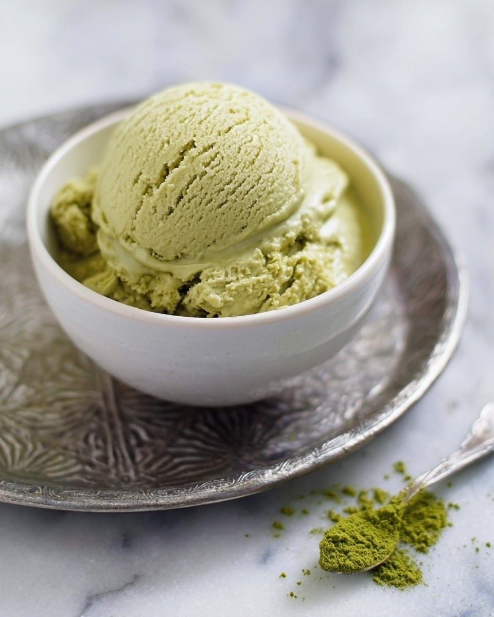 A small white bowl filled with one large scoop of pale green matcha ice cream on top of a slightly uneven layer of the same ice cream. The texture looks creamy and slightly crumbly. The bowl rests on a round silver tray with an intricate carved pattern. There is a small pile of loose matcha powder near the bowl on the white marbled surface, and a silver spoon lies next to it. Photo taken with an iphone --ar 4:5 --v 7