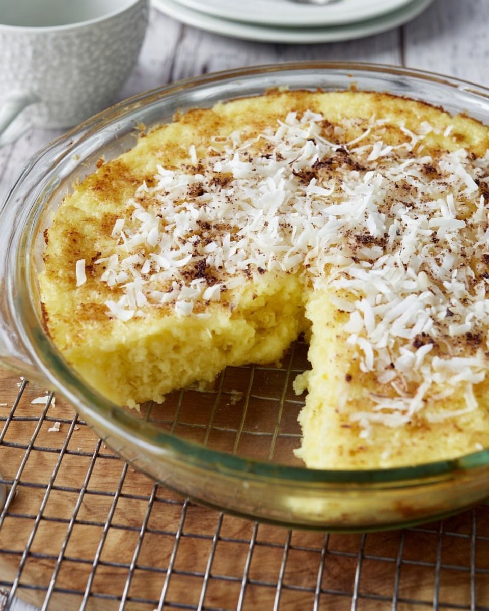A round clear glass pie dish contains a sliced yellow baked pudding with a rough soft texture, topped with white shredded coconut and small dark brown specks spread evenly on the surface. One square piece is pulled slightly out from the rest, showing the thick creamy inside with a golden crust on top. The dish is placed on a metal grid rack over a light wooden surface, and in the blurred background, there is a white cup and bowl on a white marbled texture. photo taken with an iphone --ar 4:5 --v 7