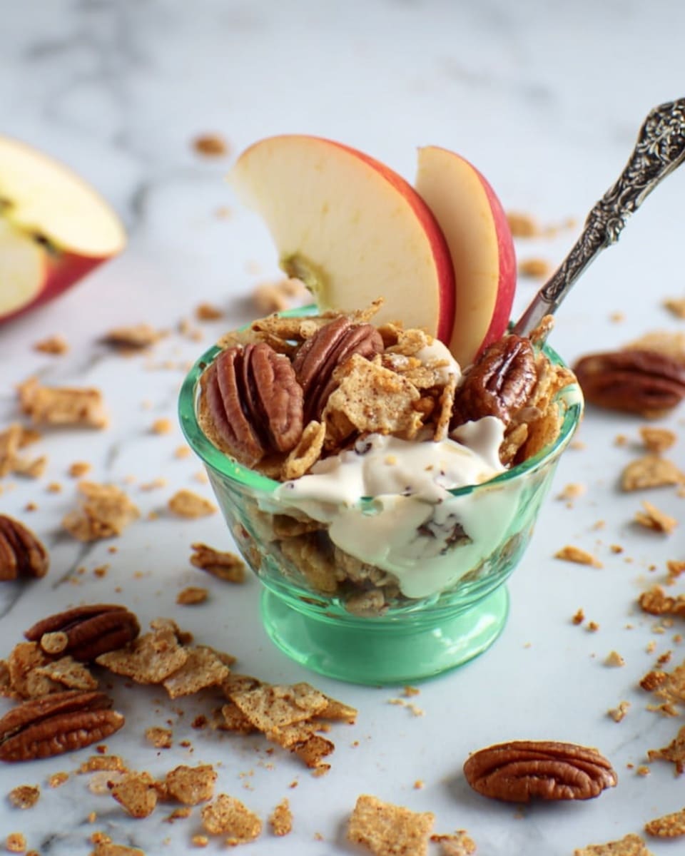 The image shows a small green glass bowl filled with layers. The bottom layer is a cluster of light brown cereal pieces with a crispy texture. On top, there are several pecan halves with a shiny, rich brown color. White creamy yogurt is drizzled over the pecans and cereal, creating a smooth, glossy contrast. Two thin, tall slices of red and cream-colored apple are placed upright at the back of the bowl. A silver spoon with a decorative handle is stuck in the bowl, partially covered with yogurt. Around the bowl on a white marbled surface, scattered cereal crumbs, pecan halves, and small pieces of apple are spread loosely. Photo taken with an iphone --ar 4:5 --v 7