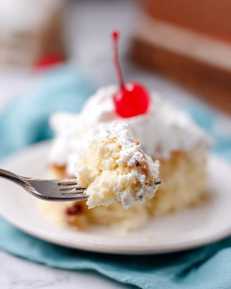 A close-up view shows a fork holding a bite of a layered dessert with a soft, light yellow cake layer mixed with small brown bits, topped with fluffy white cream. In the blurred background, a white plate holds the rest of the dessert, which appears creamy and moist, with a bright red cherry placed on top. The plate sits on a soft blue cloth over a white marbled surface. The scene has a warm and inviting feel, focused on the delicious texture of the dessert on the fork. photo taken with an iphone --ar 4:5 --v 7