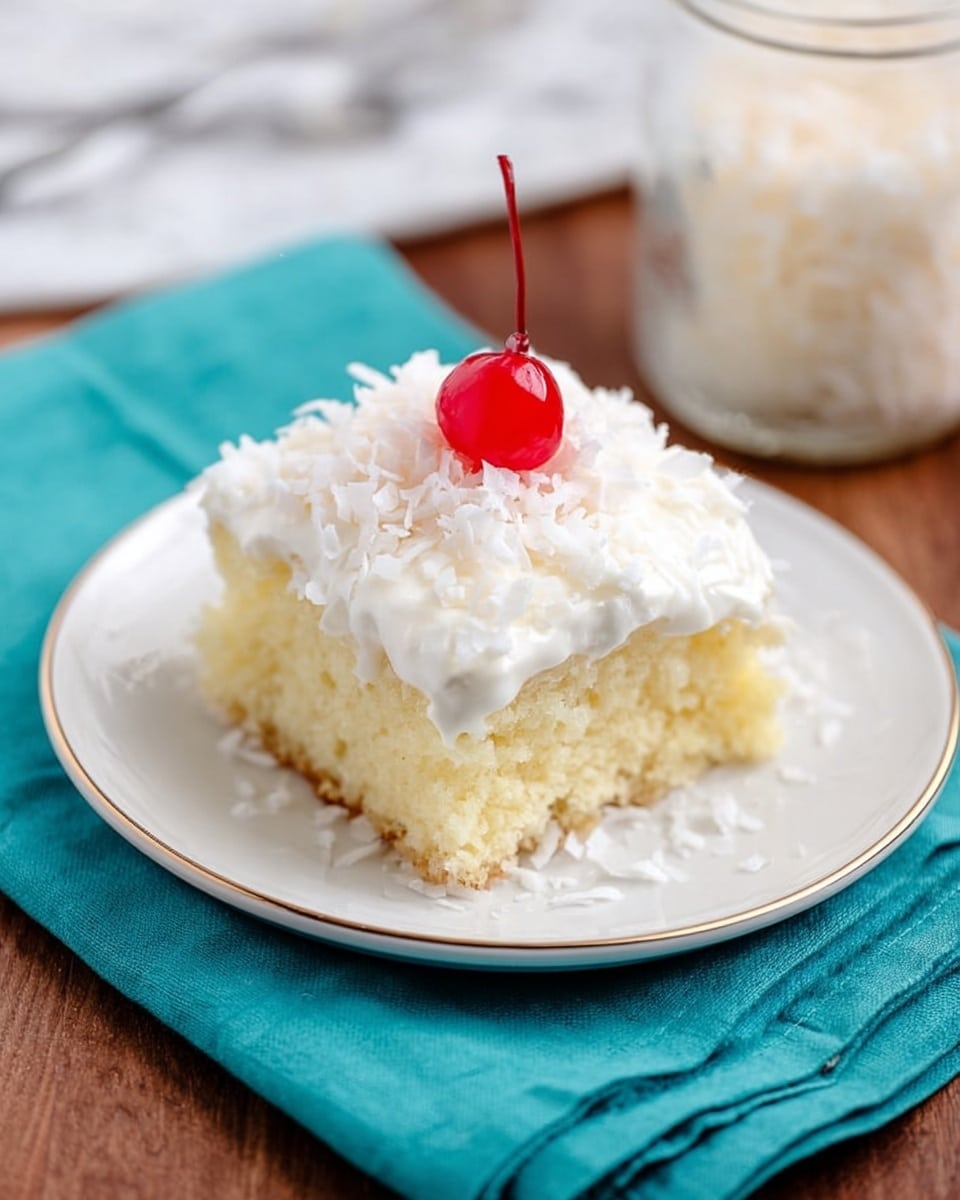 A square piece of light yellow sponge cake with a soft and moist texture sits on a white plate with a gold rim, which is placed on a teal cloth napkin over a wood surface. The cake is topped with a thick layer of white creamy icing, sprinkled with shredded coconut flakes, and finished with a single bright red cherry in the center. In the background, there is a blurred glass jar with more shredded coconut. The whole scene rests on a white marbled texture surface. photo taken with an iphone --ar 4:5 --v 7