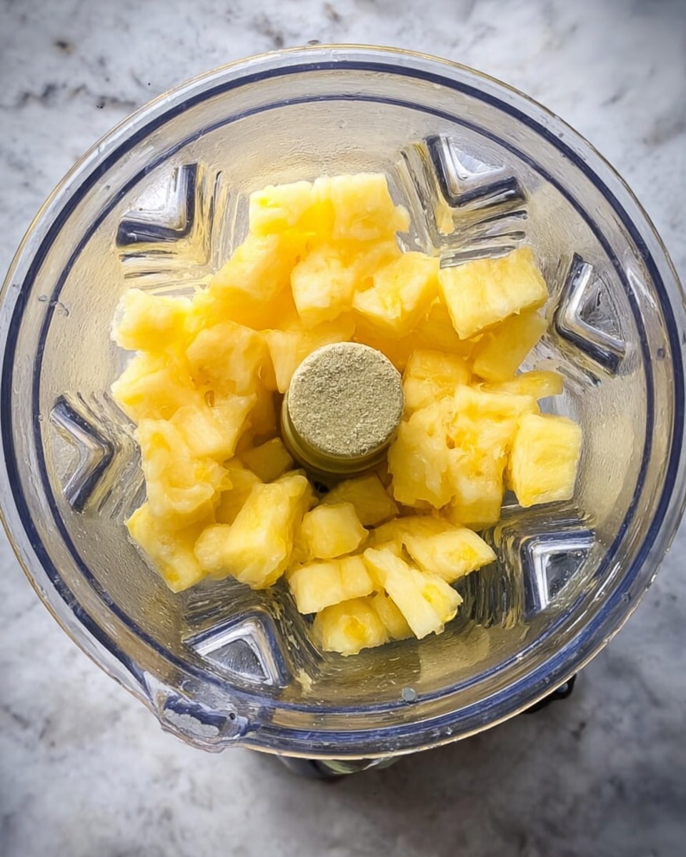 A close-up top view of a clear blender container filled with small yellow chunks of pineapple resting at the bottom center. The blender sits on a white marbled surface, and the container’s inner blades and ridges are visible, showing a shiny, textured clear plastic. The top edges of the blender and part of its base are also visible, framed by the white marbled background. photo taken with an iphone --ar 4:5 --v 7