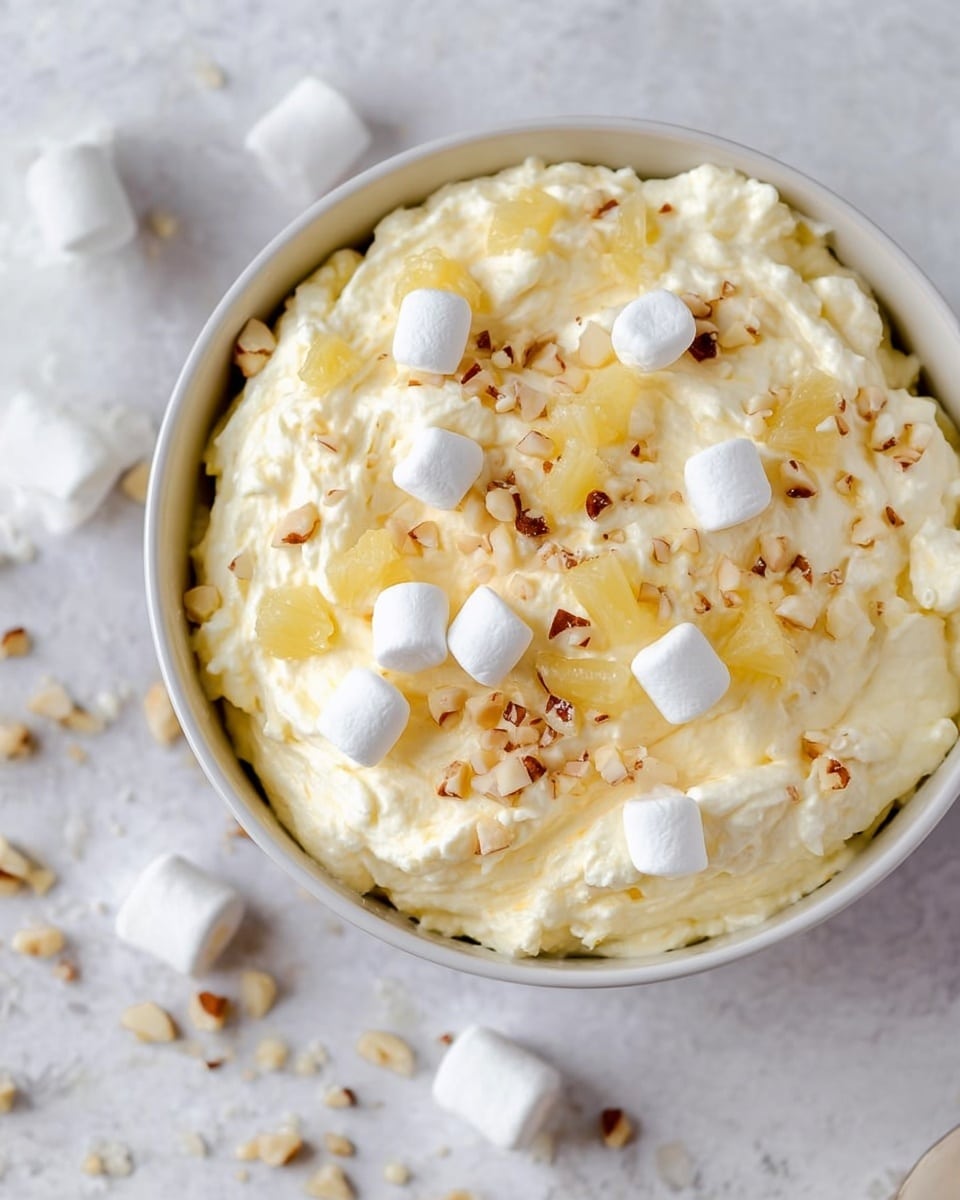 A close-up image of a white bowl filled with a creamy, pale yellow mixture that has a soft and fluffy texture. On top of the mixture, there are small white marshmallows, golden pineapple chunks, and small pieces of chopped nuts scattered evenly over the surface. The bowl is placed on a white marbled texture with a few marshmallows and nut pieces resting on the surface nearby. photo taken with an iphone --ar 4:5 --v 7