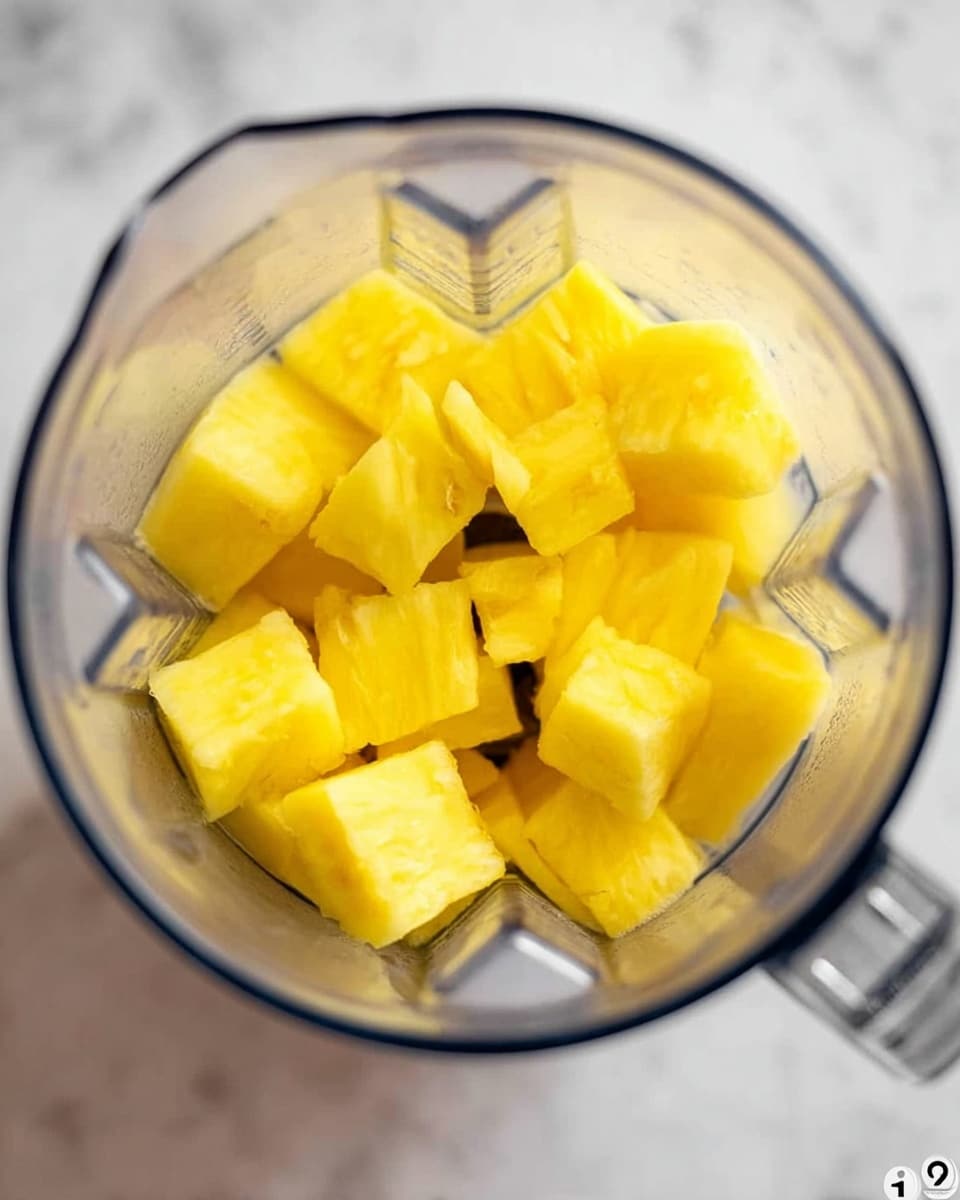 The image shows a clear blender container filled with bright yellow pineapple chunks. The pieces are thick and uneven, sitting at the bottom of the blender, and the container is on a white marbled surface. The blender blade is visible on the right inside the container. Photo taken with an iphone --ar 4:5 --v 7