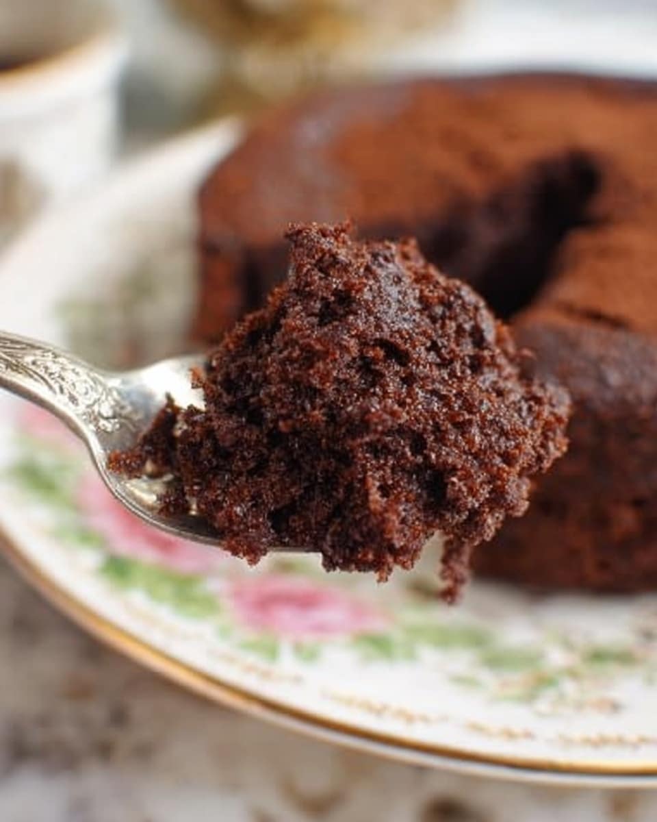 The image shows a close-up of a silver spoon holding a thick, dark brown chocolate cake piece with a rough, crumbly texture. The spoon is in focus in the middle of the frame, with the rest of the white plate with purple and green floral patterns blurred in the background. The plate holds the remaining part of the round chocolate cake, which has a dense look with a shiny surface. The scene is set on a white marbled surface. Photo taken with an iphone --ar 4:5 --v 7