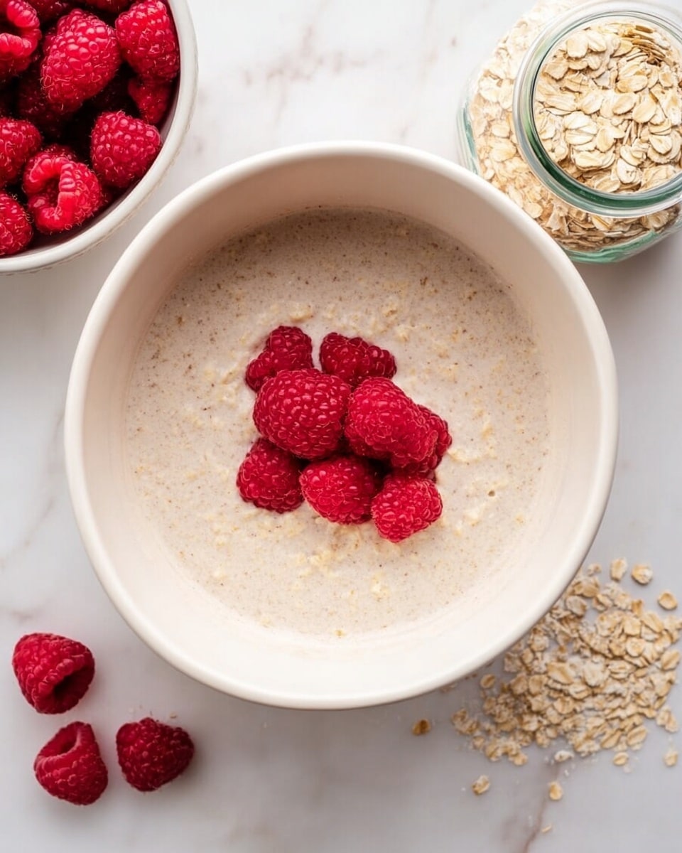 A white bowl sits on a white marbled surface, filled with a creamy oatmeal base that has a light tan color and a smooth texture. On top of the oatmeal, a small cluster of fresh, bright red raspberries is placed slightly off-center, adding a vibrant pop of color. Nearby, to the right of the bowl, is a small clear glass jar filled with dry rolled oats, beige and flaky in texture. To the left of the bowl, a white bowl holds more fresh raspberries, and a few extra raspberries are scattered casually on the white marbled surface around it. photo taken with an iphone --ar 4:5 --v 7