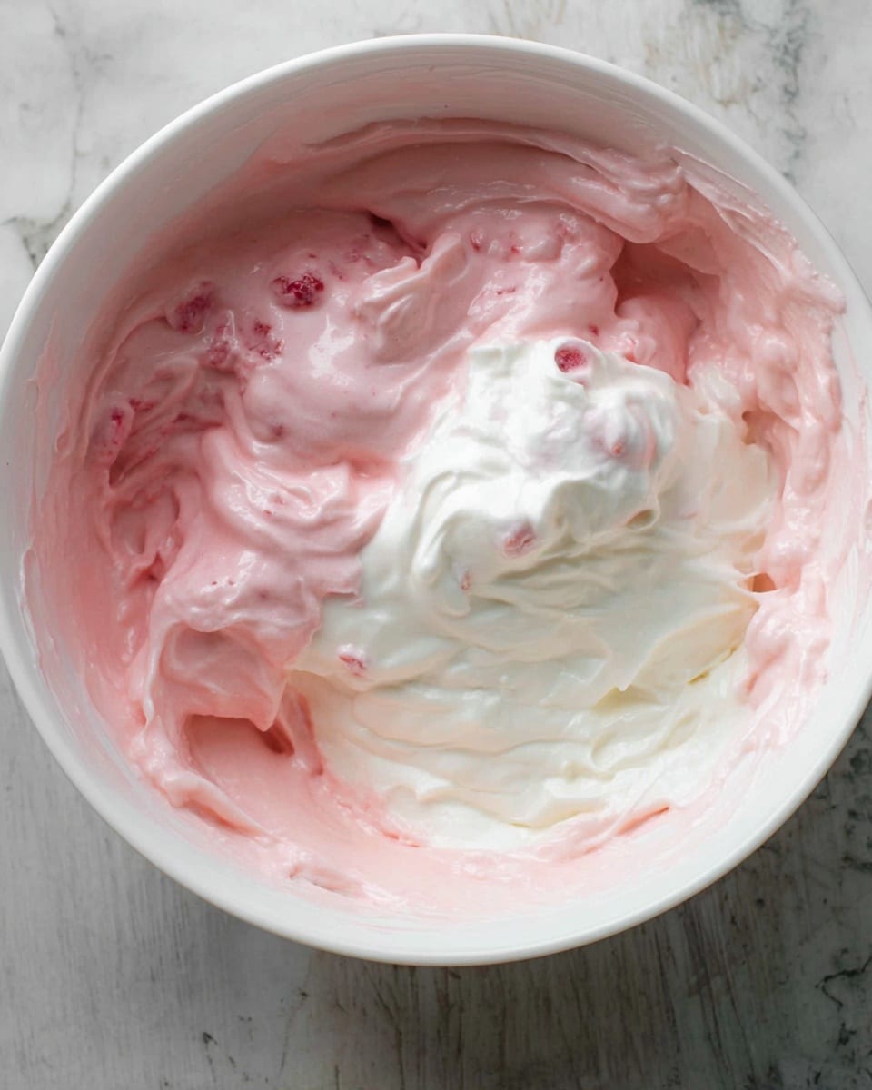 In a white bowl, there is a mix of two creamy layers: one is smooth and pink with small fruit pieces, and the other is thick and white, swirled together but still partly separate. The pink layer covers most of the bottom and sides of the bowl with some patches of white cream on top, creating a marbled effect. The textures look soft and slightly fluffy, showing folds and peaks. The bowl rests on a white marbled surface. Photo taken with an iphone --ar 4:5 --v 7