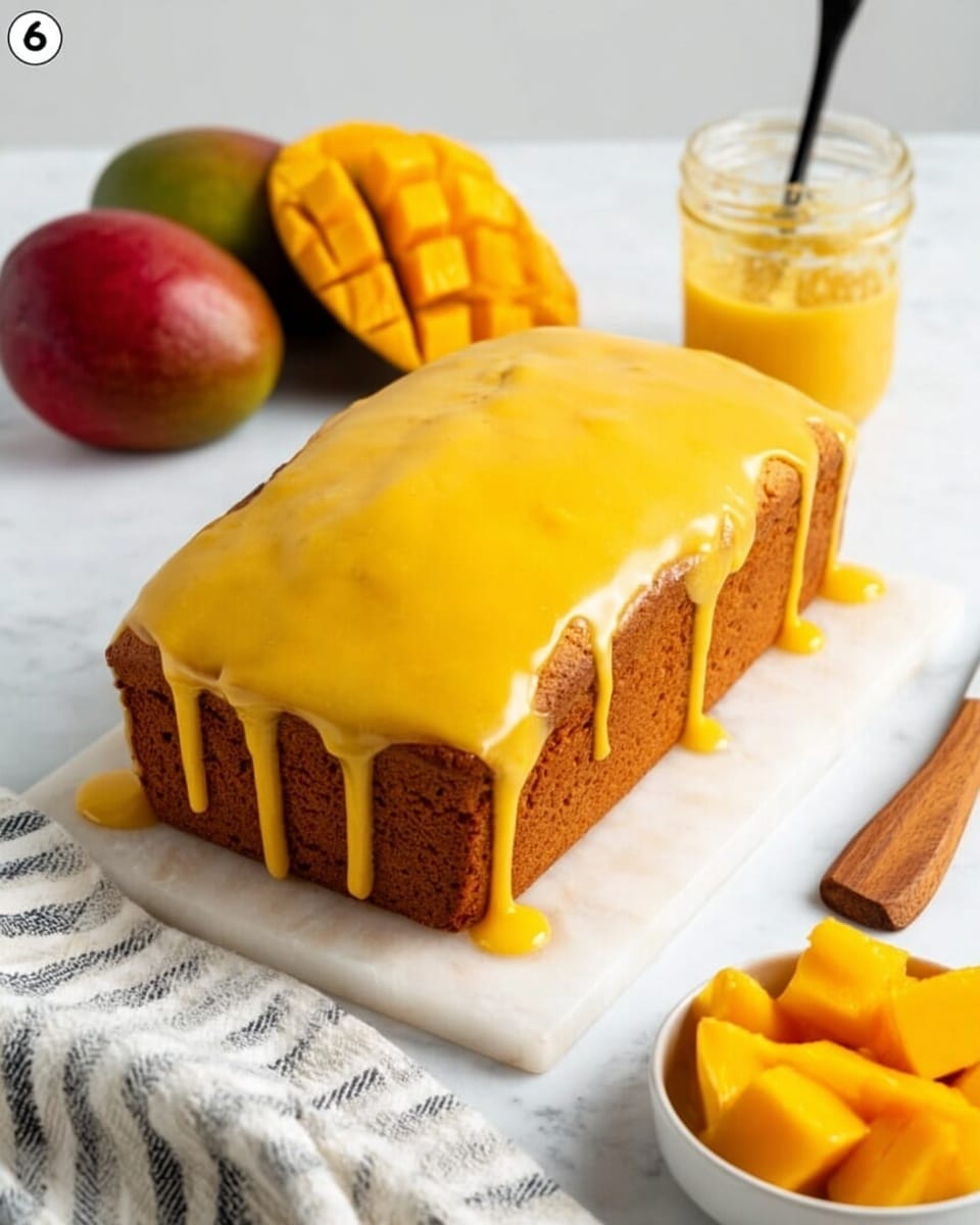 A loaf cake sits on a white surface with a white marbled texture, covered with smooth, bright yellow glaze that drips down the sides, creating a shiny and soft look. The cake itself is light brown with a soft crumb texture visible where the glaze does not cover. In the background on the left, there is a whole mango with a dark reddish-yellow skin and a mango half with bright orange flesh cut into cubes. On the right side, a small clear glass jar holds thick yellow sauce with a black spoon inside. A wooden spatula lies nearby, and on the bottom right, a small white bowl contains chunks of bright yellow mango. A striped cloth is placed on the surface near the bottom left corner. Photo taken with an iphone --ar 4:5 --v 7