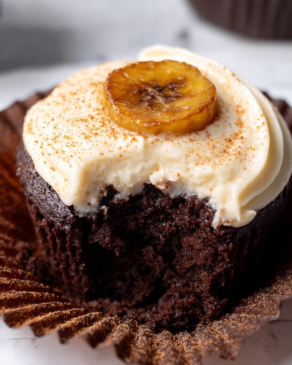 The image shows a close-up of a single dark brown chocolate cupcake with moist texture, in a opened dark brown paper liner resting on a white marbled surface. The cupcake has one thick layer of smooth creamy white frosting on top, sprinkled lightly with cinnamon powder. On the center of the frosting sits one caramelized golden-brown banana slice. The cupcake looks soft and rich, with a slightly rough cake crumb visible where a bite has been taken. Photo taken with an iphone --ar 4:5 --v 7