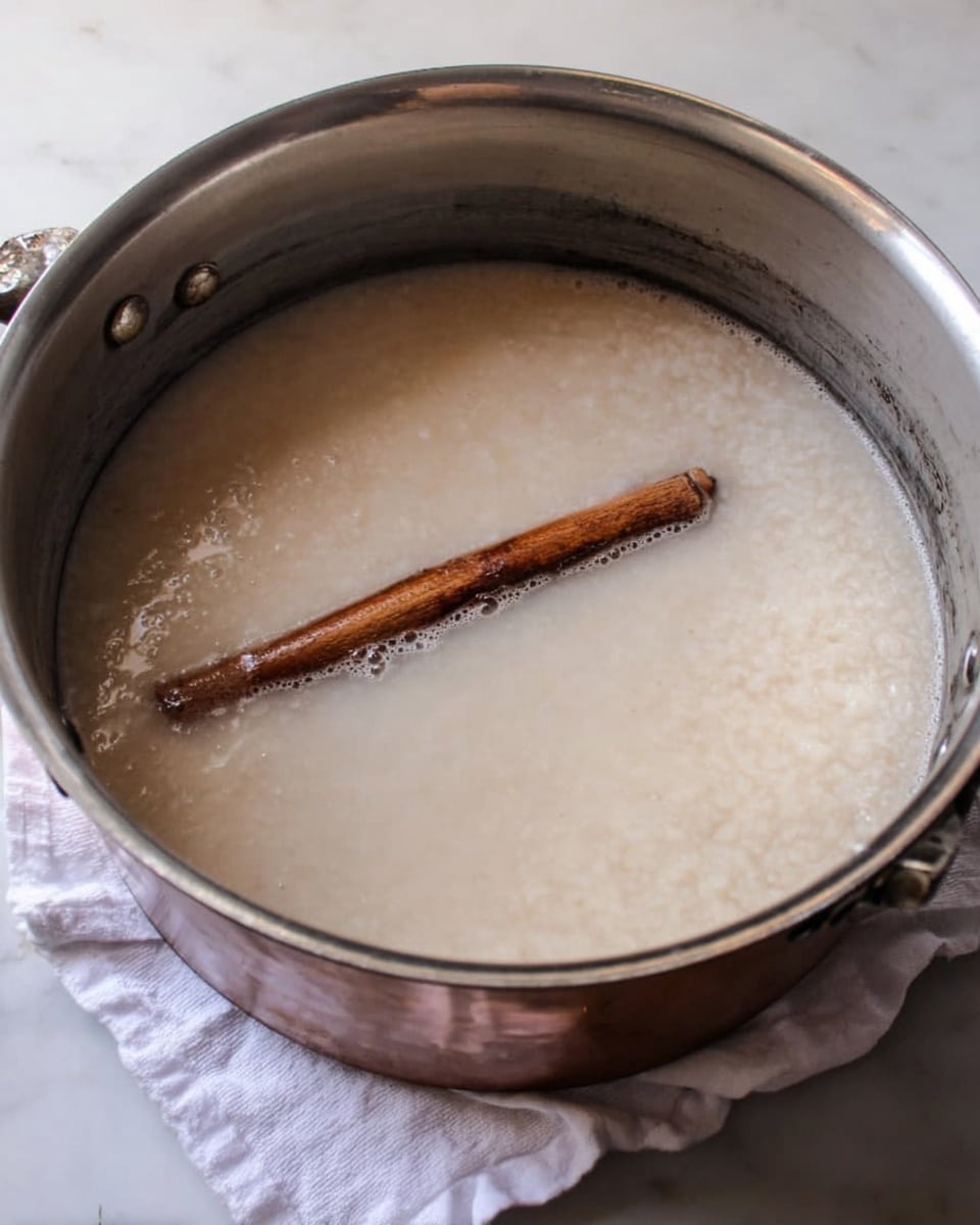 A large silver pot filled with a thick, off-white liquid that looks creamy and slightly lumpy. Inside the liquid, there is a single brown cinnamon stick partially submerged near the center, adding contrast with its dark, rough texture. The pot is placed on a white marbled surface, and a white cloth is seen underneath or beside it. The inside walls of the pot are shiny and metallic, showing signs of use. Photo taken with an iphone --ar 4:5 --v 7