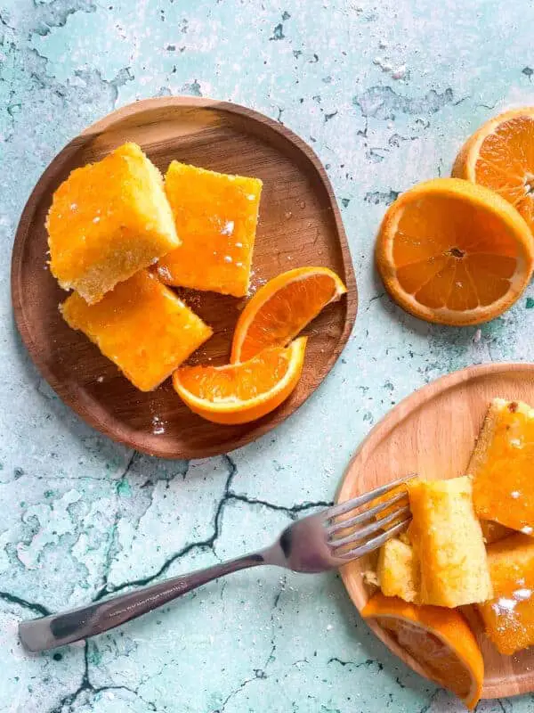 The image shows two wooden round plates on a white marbled surface. The plate at the top left holds three square yellow pieces of cake with a shiny glaze on top. Beside the cake pieces are two orange wedges with bright, juicy flesh. The plate at the bottom right contains three square yellow cake pieces and one piece that has been split apart, showing its soft texture inside. There is a silver fork resting diagonally across this plate, touching the split cake piece. Next to this plate are two orange halves with vibrant orange insides. The photo taken with an iphone --ar 4:5 --v 7