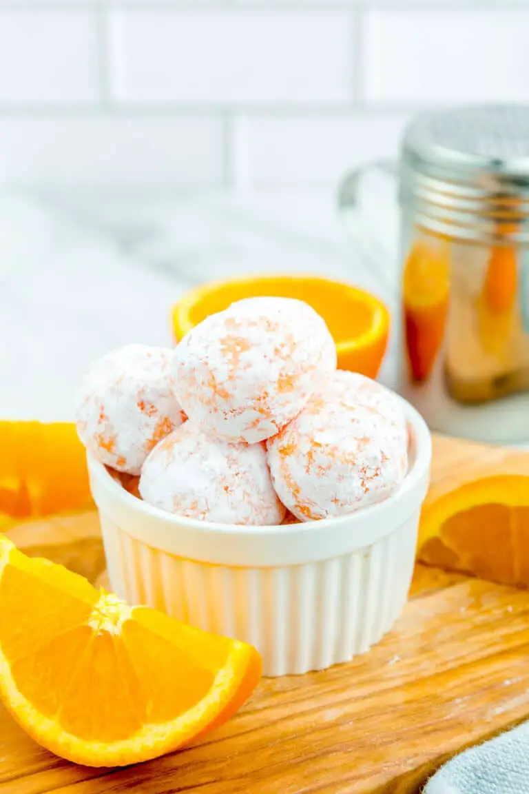 A small white ramekin filled with five round, orange-colored cookies covered lightly in white powdered sugar sits on a wooden board. The cookies have a cracked texture showing glimpses of the orange color underneath. Around the ramekin, there are fresh bright orange slices placed on the wooden board. In the blurred background, there is a silver shaker and a white marbled surface with white tiles. The scene is bright and clean, emphasizing the contrast between the white powdered sugar and the orange tones of the cookies and fruit photo taken with an iphone --ar 4:5 --v 7
