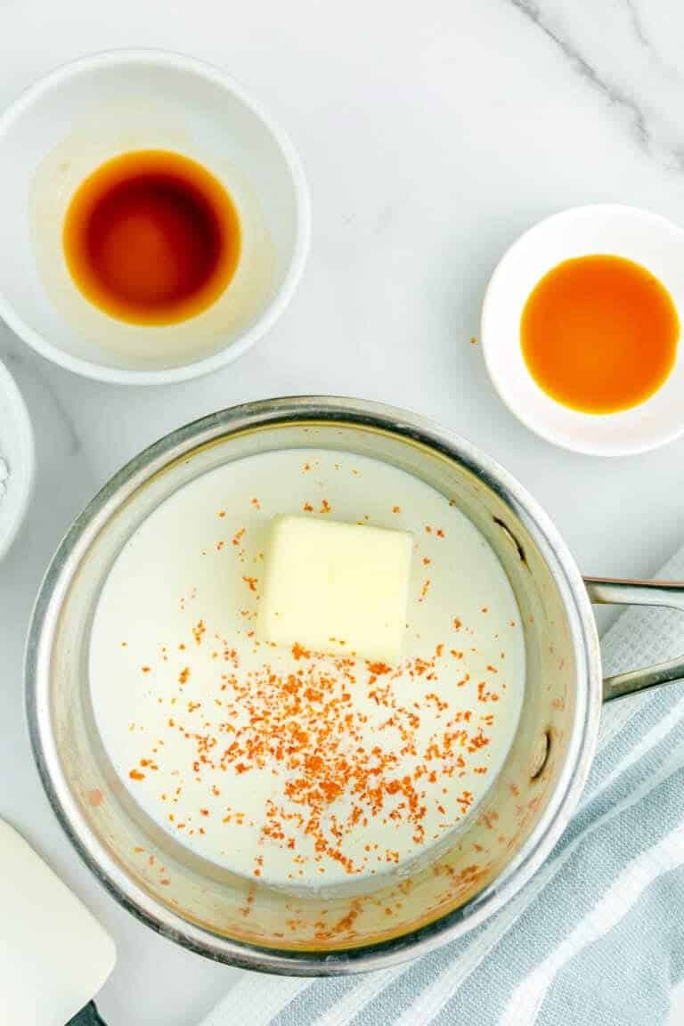 A metal saucepan filled with a white liquid base, topped with a square piece of pale yellow butter and small orange specks lightly scattered across the surface. The pan is placed on a white marbled surface with a soft blue and white striped cloth underneath. Around the saucepan, there are three small white bowls containing different liquids: one with a dark brown liquid, one with a bright orange liquid, and the third is mostly out of view but partially holding a white ingredient. The scene is clean and bright with natural lighting. photo taken with an iphone --ar 4:5 --v 7
