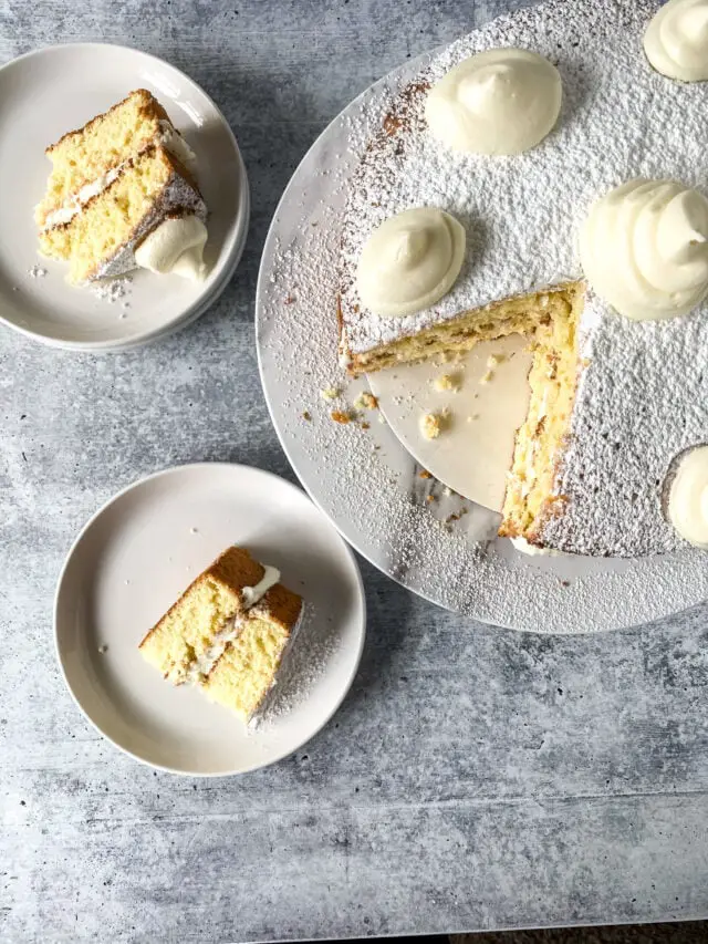 A round cake with three visible layers of light yellow sponge and white cream between each layer sits on a white plate with powdered sugar dusted over the top and five large dollops of white cream spaced around the edge. Two slices of the same cake are served on separate white plates nearby, each slice showing the same three layers and cream filling, with one slice topped by a swirl of white cream. The cakes and plates rest on a white marbled surface. Photo taken with an iphone --ar 4:5 --v 7