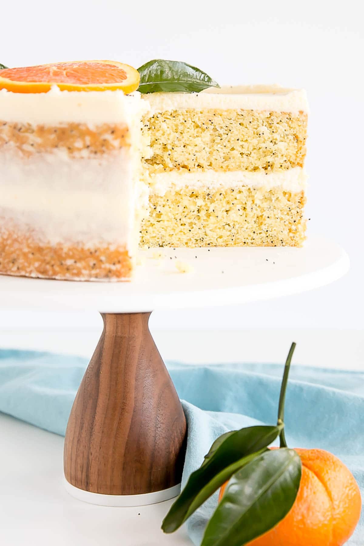 A two-layer yellow cake with small black seeds inside is shown on a white cake stand with a wooden base. Each cake layer is separated by a thick white cream layer, and the outside is thinly covered with the same white cream, letting the cake texture show through. On top, there is a round orange slice and two green leaves as decoration. The background and surface are white with a marbled texture, and there is a light blue cloth and a fresh orange with green leaves placed near the stand. Photo taken with an iphone --ar 4:5 --v 7