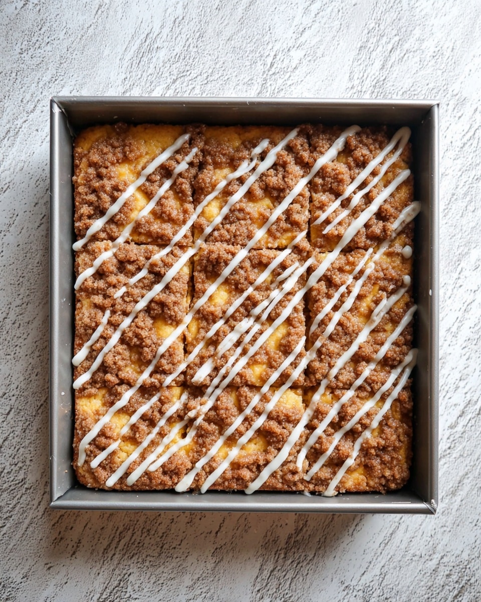 A square baking pan filled with a thick, golden-yellow cake base as the bottom layer, topped with a textured layer of crumbly brown cinnamon sugar streusel spread unevenly across the surface, and finished with a thin drizzle of white icing running diagonally in parallel lines over the streusel. The pan is on a white marbled textured surface. photo taken with an iphone --ar 4:5 --v 7
