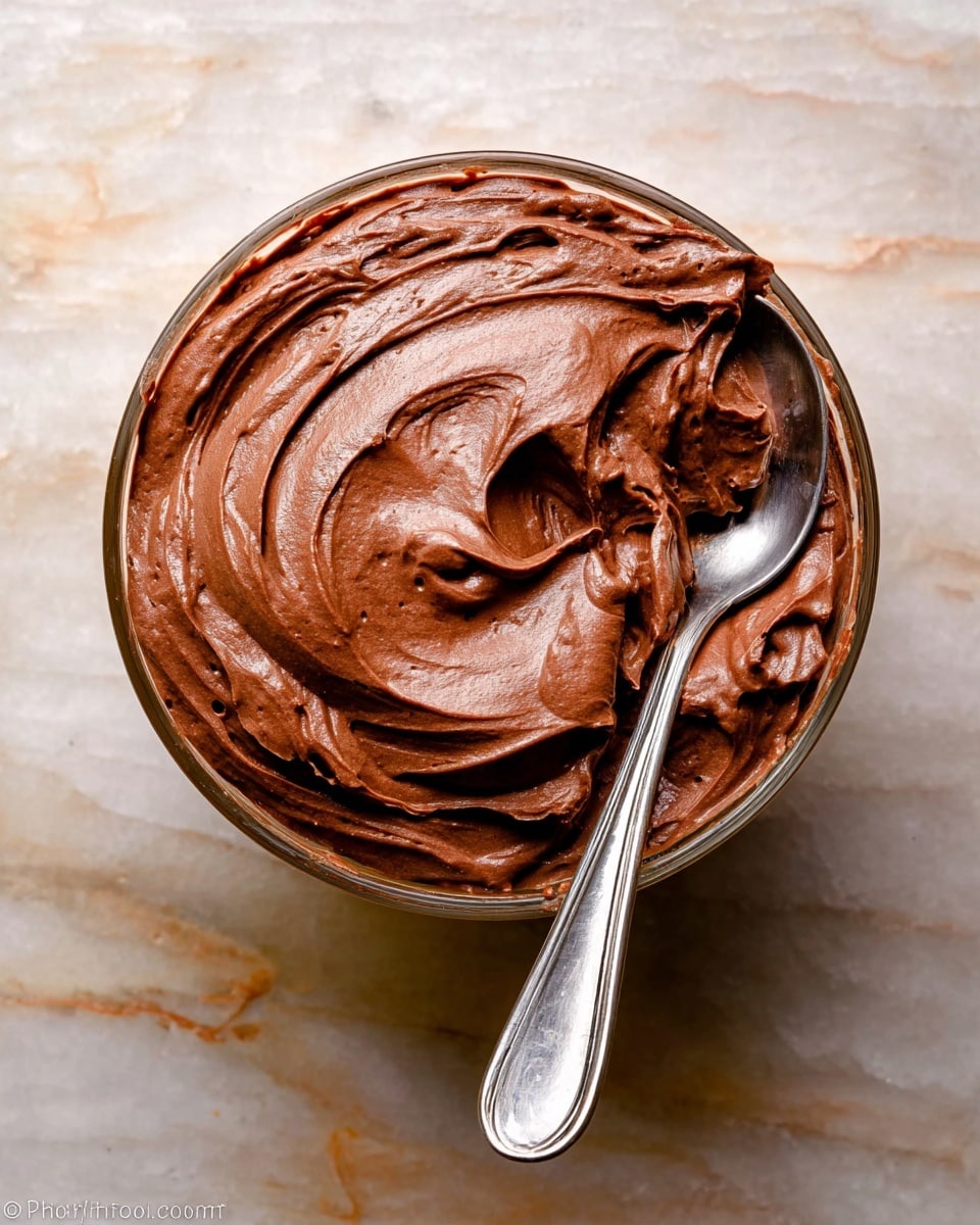 A small clear glass bowl filled with one thick layer of smooth, rich chocolate mousse, swirled on top to create soft waves and peaks. A silver spoon is partially dipped into the mousse on the right side, showing more of the chocolate texture. The bowl is on a white marbled surface. photo taken with an iphone --ar 4:5 --v 7
