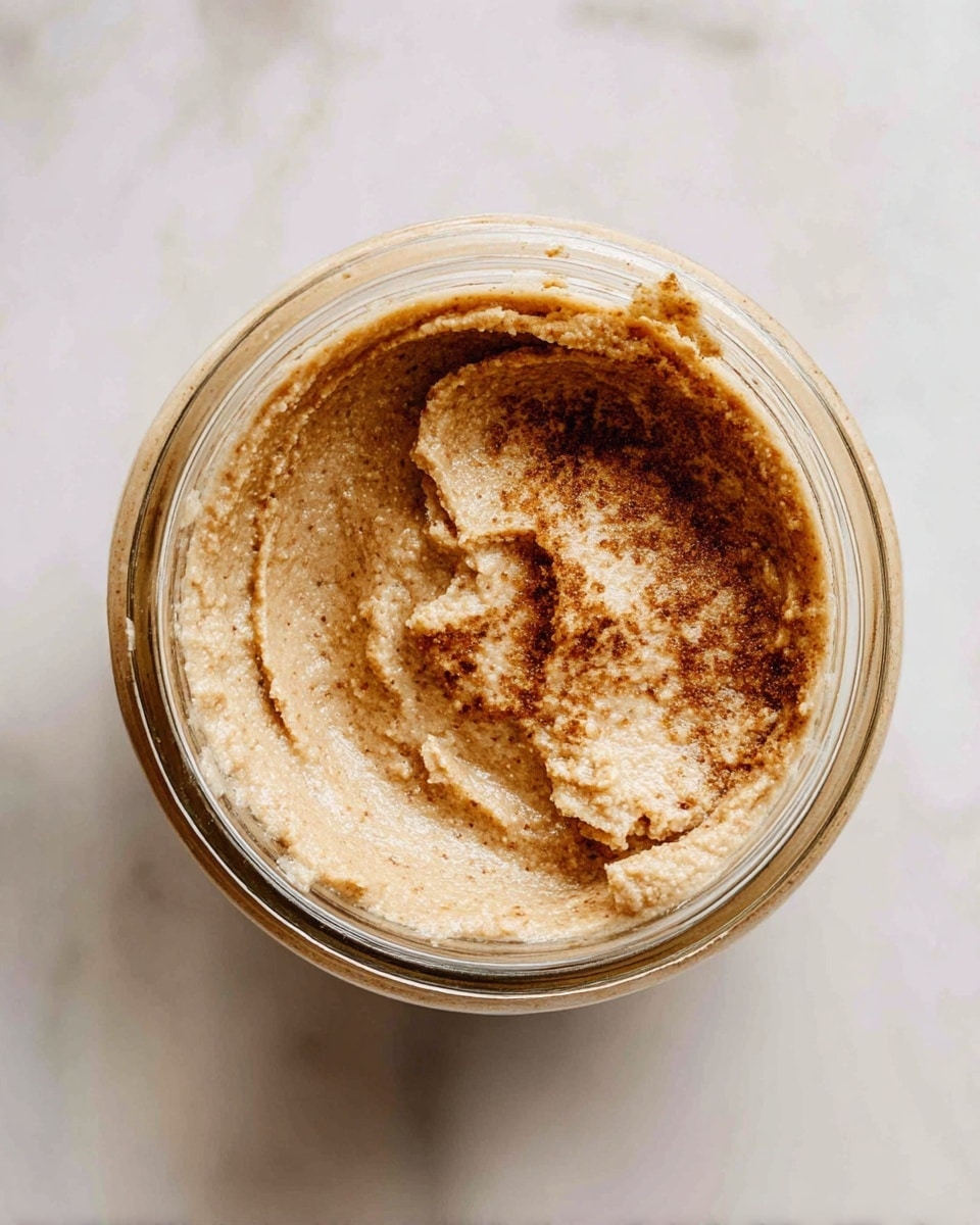 A close-up top view of a glass jar filled with a thick, creamy paste that is light brown with darker brown spots. The texture looks smooth with some small grainy bits. The paste is spread evenly inside the jar, showing slight swirls and a rough edge near the opening. The jar sits on a white marbled surface. photo taken with an iphone --ar 4:5 --v 7