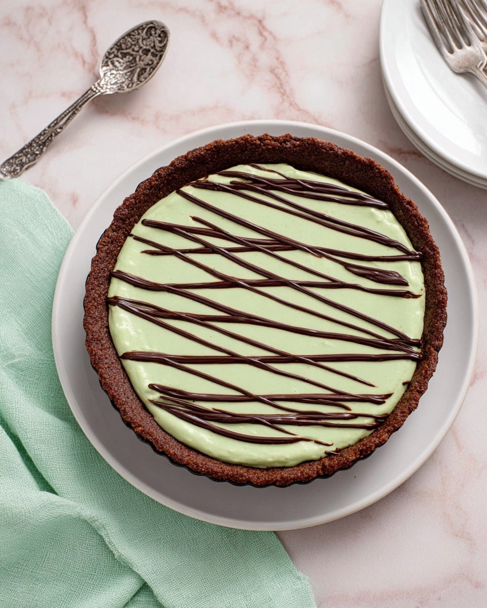 A round tart with a thick, dark brown crust forming the base and edges. Inside the crust is a smooth, light green creamy layer spread evenly in a circular shape. Over this green layer, there are thin, dark chocolate drizzles crossing diagonally from top left to bottom right in irregular lines. The tart sits on a white plate placed on a white marbled surface. Nearby, there is a silver decorative spoon on a light green cloth to the left and a small white plate with three silver forks on the right. photo taken with an iphone --ar 4:5 --v 7
