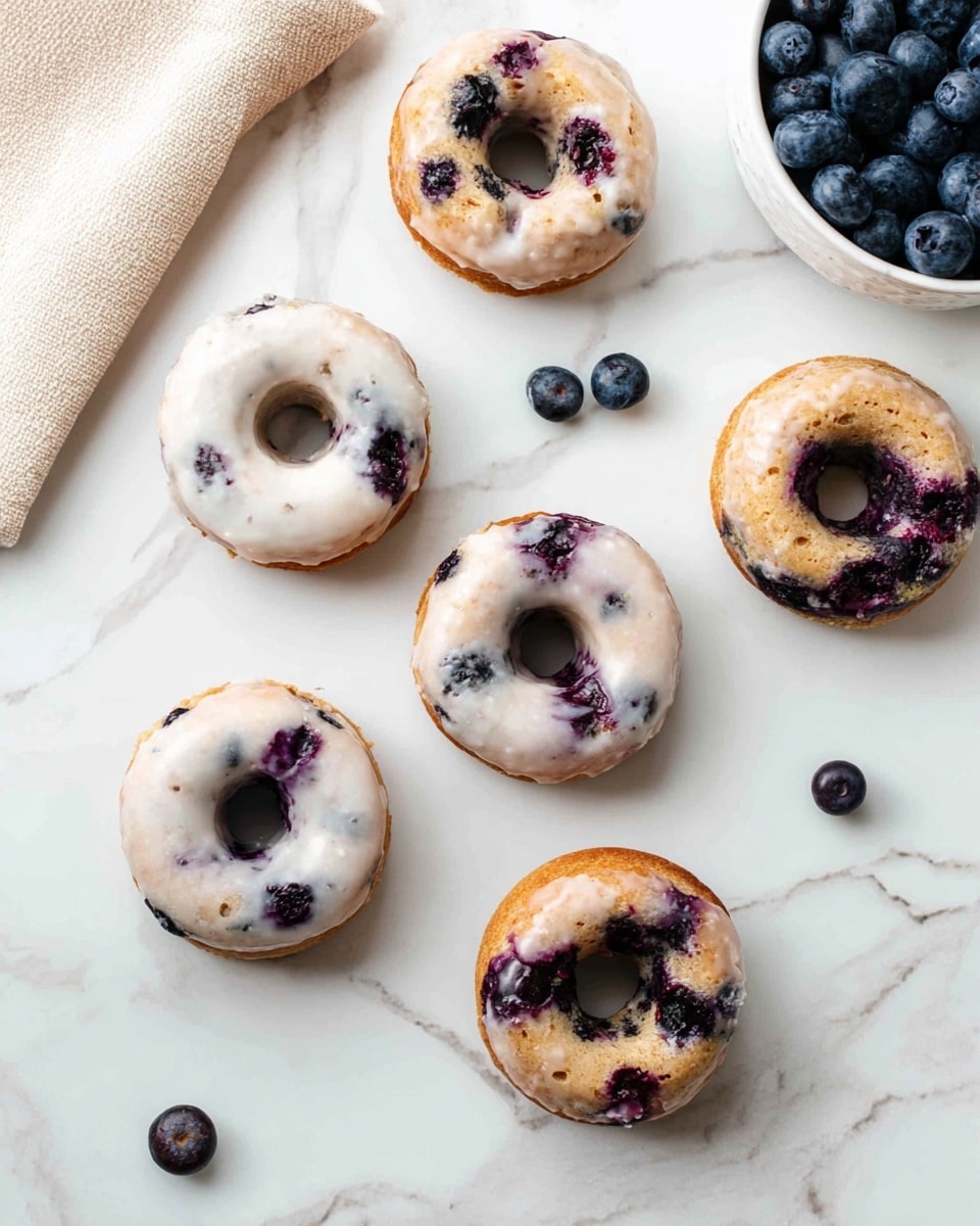 The image shows six small blueberry donuts placed on a white marbled surface. Three of them have a smooth white glaze covering the top, with the glaze slightly melting and allowing some blueberry spots to show through. The other three donuts have a golden-brown baked texture with visible dark purple blueberry spots inside the dough. Scattered around the donuts are a few whole blueberries, and in the top right corner, there is a white bowl filled with more blueberries. A beige cloth is partially visible in the top left corner. photo taken with an iphone --ar 4:5 --v 7
