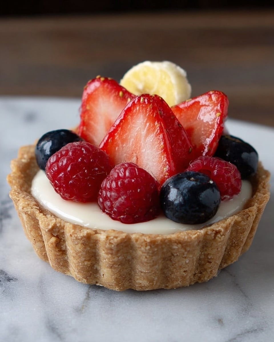 A small tart with a light brown, crumbly crust forms the base. Inside, a creamy white layer fills the crust. On top of this are several pieces of fresh fruit: thin, fan-shaped slices of red strawberries sit in the center, surrounded by whole shiny red raspberries and dark blue blueberries. A small yellow banana piece peaks out from the back of the fruit topping. The tart rests on a white marbled surface. Photo taken with an iphone --ar 4:5 --v 7