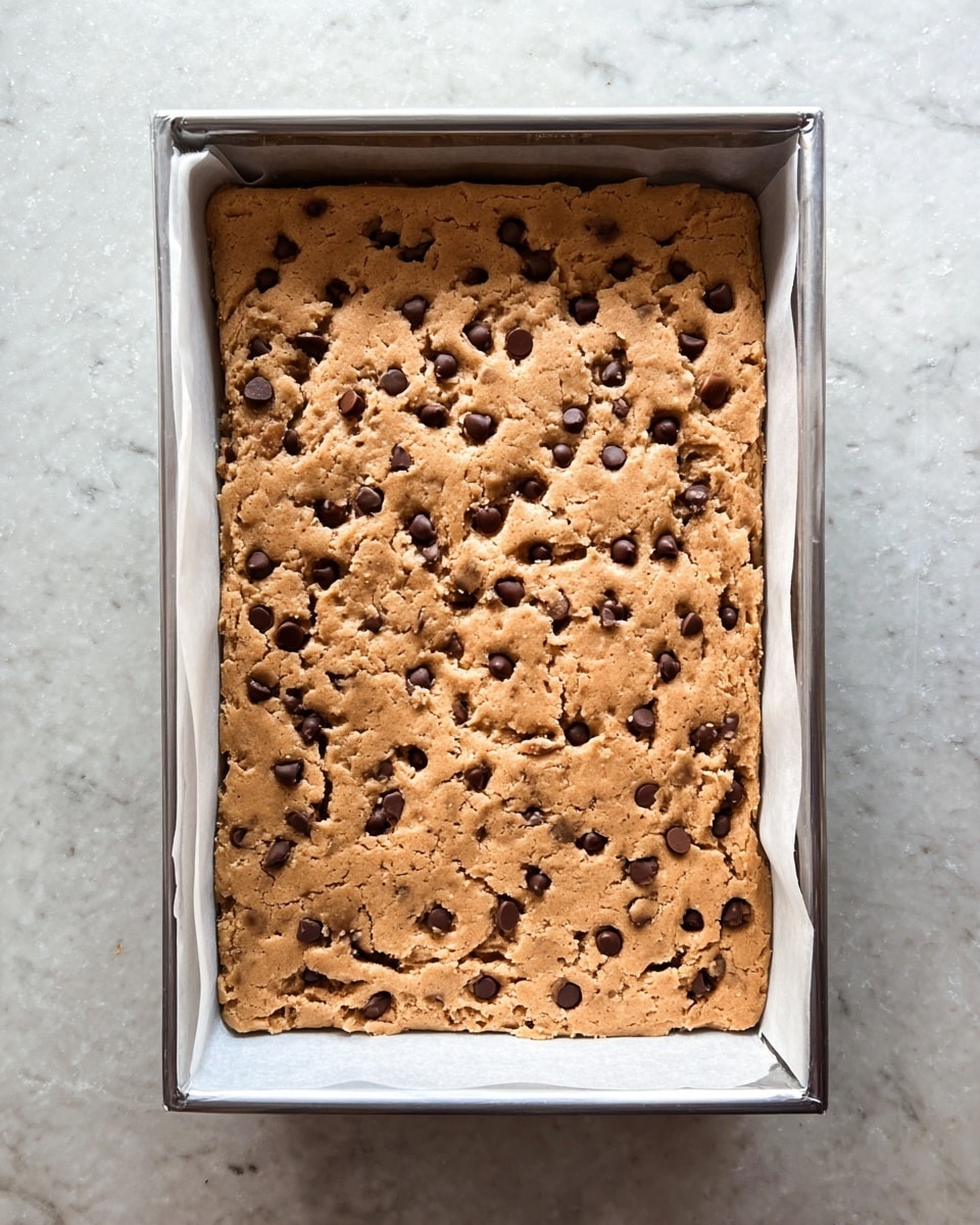 A single thick layer of golden brown cookie dough with a slightly rough texture fills a rectangular metal baking pan lined with white parchment paper. The cookie dough surface is scattered with semi-melted dark chocolate chips, some slightly sunk into the dough and others resting on top. The edges of the dough are slightly raised and cracked, showing a soft, chewy inside. The pan sits on a white marbled surface, and the image shows the entire pan from above. photo taken with an iphone --ar 4:5 --v 7