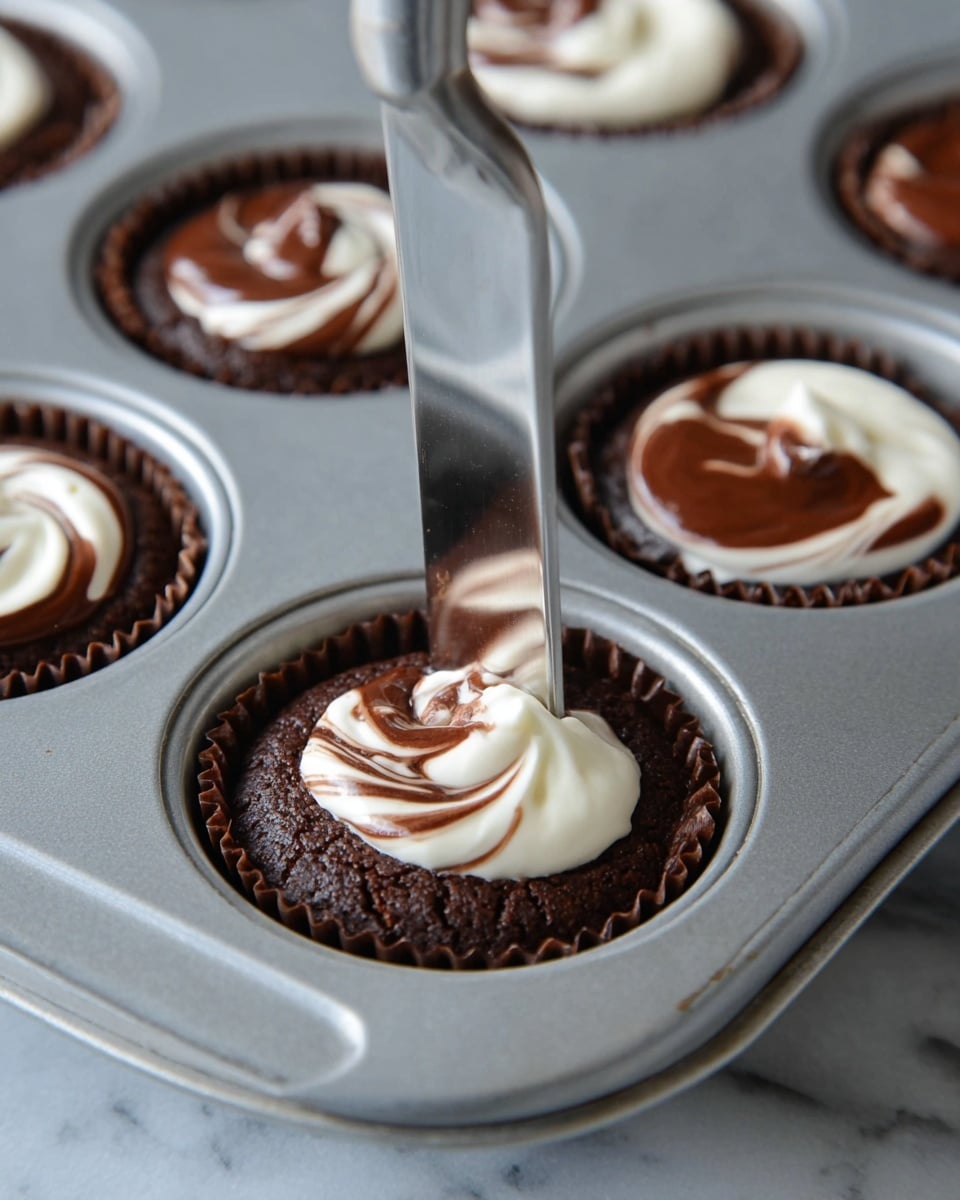 A close-up of several cupcake liners filled with dark brown chocolate batter, each topped with white cream swirled on top to create a marbled design. A shiny metal knife is gently swirling the cream and chocolate in one liner to mix the two layers smoothly. The cupcake liners are inside a metal muffin tray, placed on a white marbled surface. Photo taken with an iphone --ar 4:5 --v 7