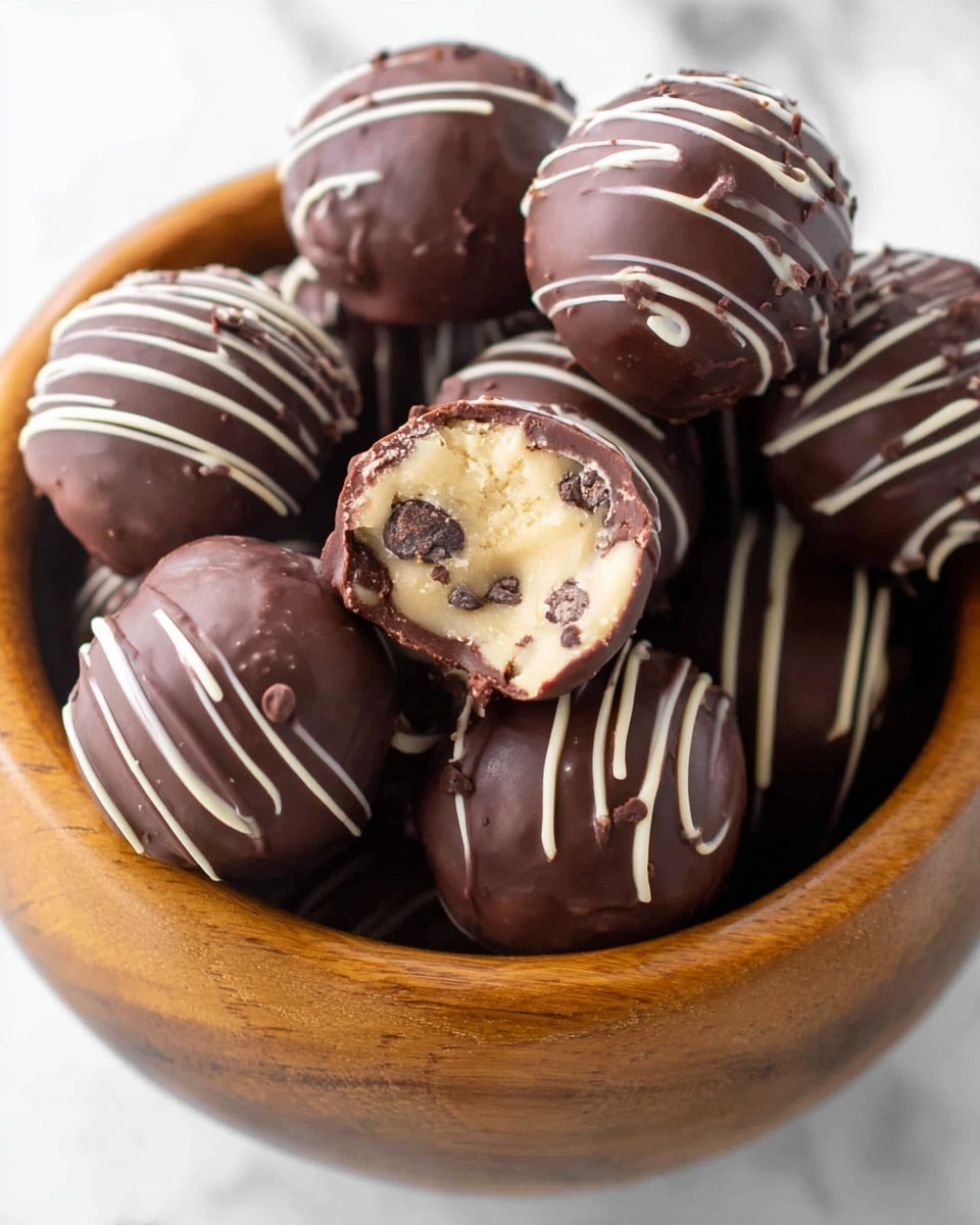 A wooden bowl filled with round chocolate balls, each coated in smooth dark chocolate; some have thin white chocolate lines drizzled on top in curved patterns. One chocolate ball is cut open to show a creamy, light beige inside with small dark chocolate chips scattered throughout its soft filling. The chocolates have shiny surfaces with slight texture from the drizzle, and the bowl sits on a white marbled surface. Photo taken with an iphone --ar 4:5 --v 7