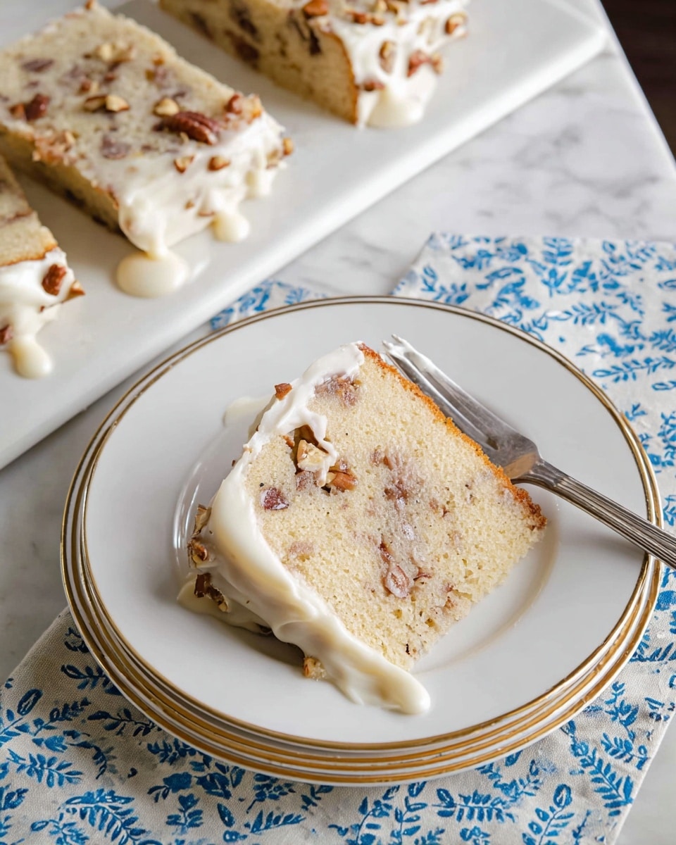 A slice of light golden cake with visible brown nut pieces is placed on a white plate with faint gold rings. The cake has a thick white frosting layer on the top, which is slightly melting and drips down the sides. The texture of the cake looks soft and moist with nuts spread inside. A silver fork rests on the plate beside the slice. Below, part of a white rectangular serving plate shows more slices of the same cake topped with chopped nuts. The setup is on a white marbled surface with a cloth featuring blue leaf patterns underneath the plates. Photo taken with an iphone --ar 4:5 --v 7