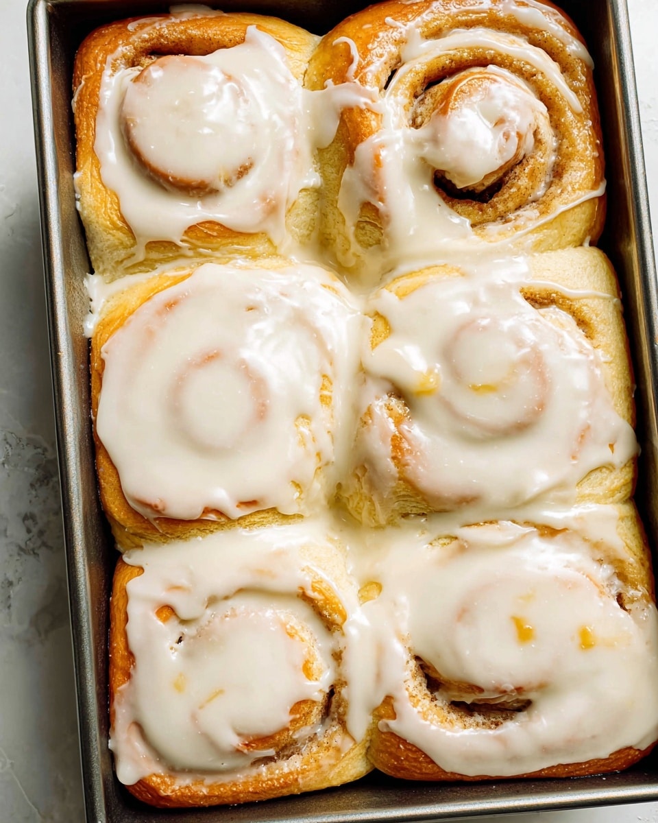 The image shows a metal pan filled with six cinnamon rolls, arranged in two rows of three. Each roll has a golden-brown base with visible swirls and a soft, fluffy texture. The top layer is thickly covered with creamy white icing that spreads unevenly, dripping slightly over the edges of some rolls, creating a smooth and shiny surface. The pan is placed on a white marbled texture background. photo taken with an iphone --ar 4:5 --v 7