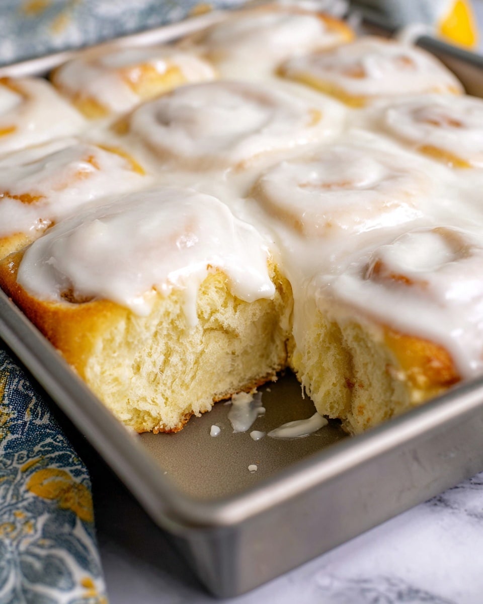 A close-up view of a metal baking pan containing soft, light golden rolls arranged in a single layer, each roll topped with a thick layer of smooth, white icing that drips slightly into the gaps between the rolls. One roll has been removed, showing the fluffy, airy texture inside with a moist crumb. The pan sits on a white marbled surface with a patterned cloth partially visible on one side. photo taken with an iphone --ar 4:5 --v 7