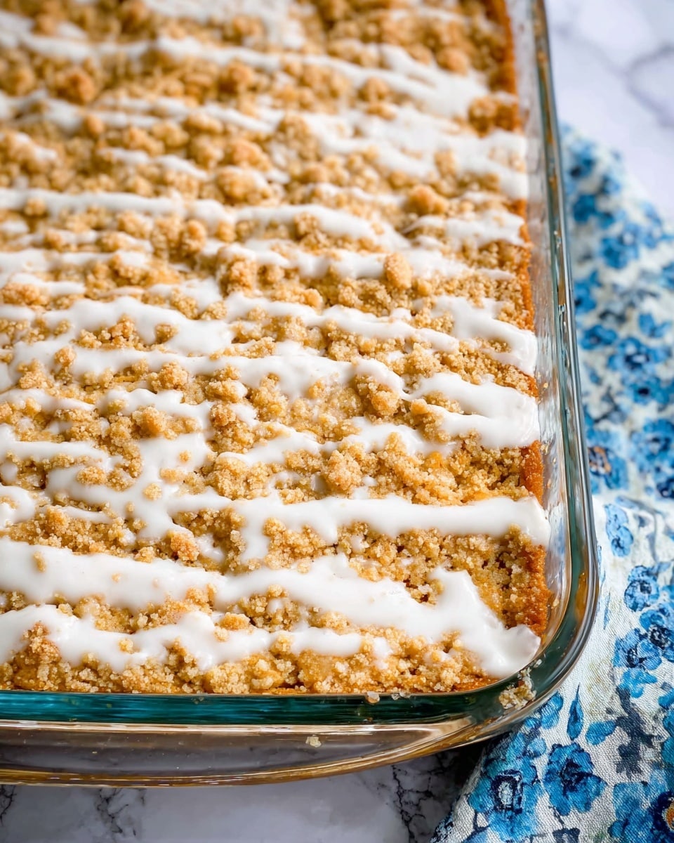 The image shows a clear glass baking dish filled with a dessert that has two main layers visible from the top: a golden brown crumbly layer covering the surface and a generous drizzle of white icing forming irregular lines across the crumb layer. The crumb topping has a rough, textured look with small clumps spread evenly, while the white icing adds a smooth, shiny contrast. The dish is set on a white marbled surface with a soft blue floral patterned fabric slightly visible on the side. The photo taken with an iphone --ar 4:5 --v 7