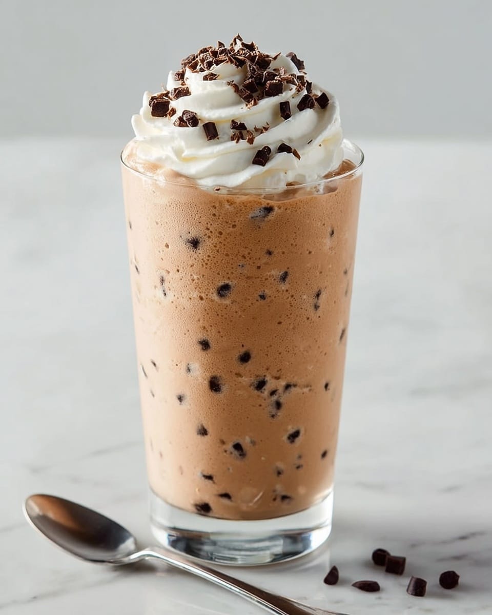 A tall clear glass filled with a creamy light brown mousse mixed with small dark chocolate chunks evenly spread inside. On top, there is a thick swirl of white whipped cream sprinkled with dark chocolate shavings. The glass is placed on a white marbled surface with a silver spoon lying next to it. photo taken with an iphone --ar 4:5 --v 7