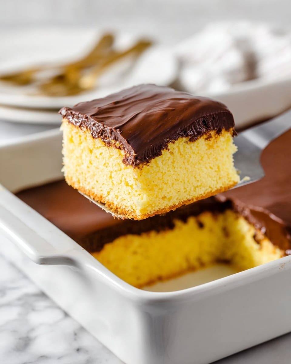 A square piece of yellow sponge cake with a soft texture has a thick layer of smooth dark brown chocolate on top. The cake piece is lifted by a metal spatula from a white rectangular baking dish filled with more of the same cake topped with an even layer of chocolate. The background shows a white marbled surface with a blurred white and gold fork on the side. Photo taken with an iphone --ar 4:5 --v 7