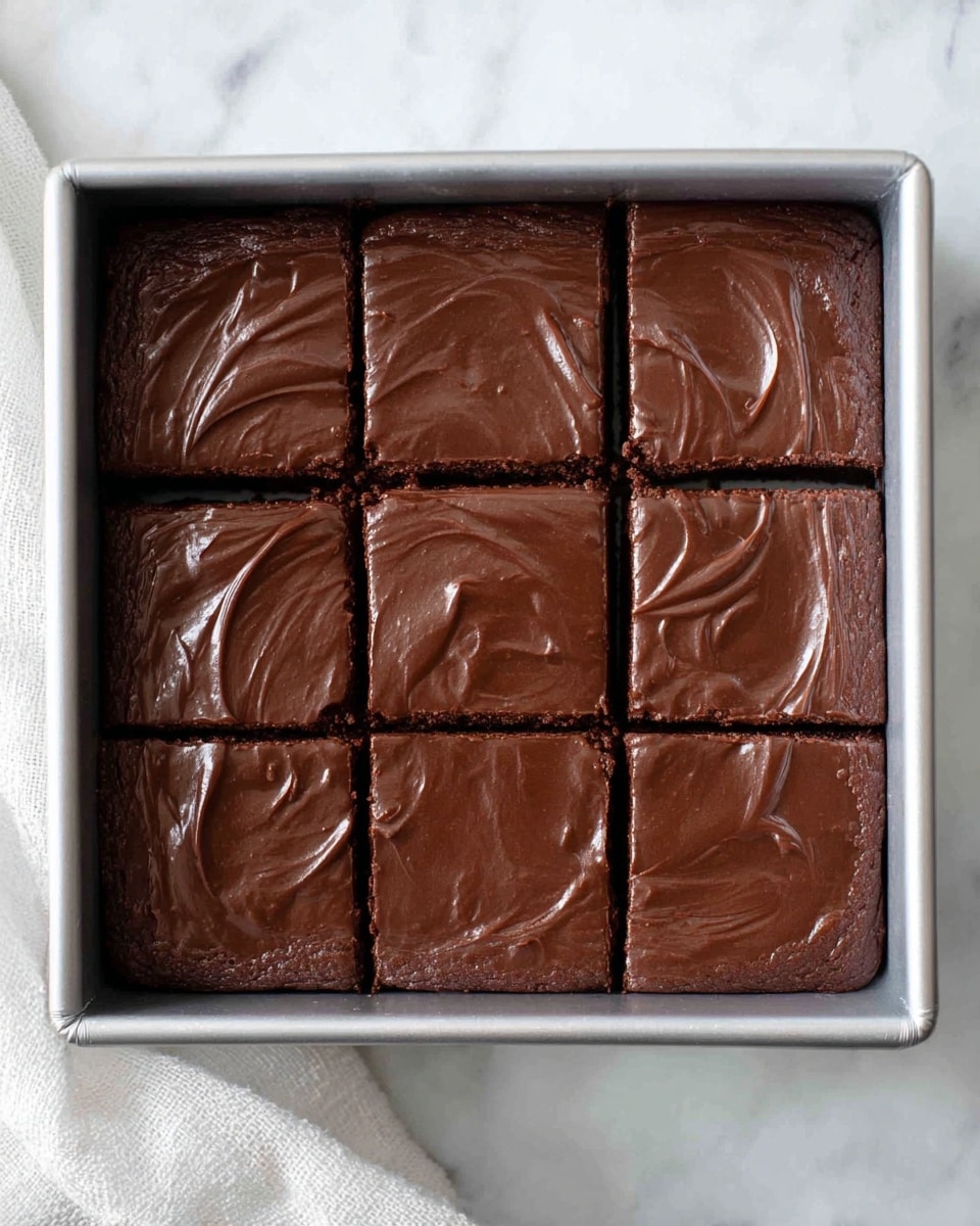 A square metal baking pan holds a thick layer of rich, smooth, dark brown chocolate batter, divided into nine even squares by shallow cuts. The chocolate surface is shiny and creamy, with subtle swirls and gentle peaks reflecting light. The pan rests on a white marbled surface with a white cloth partially visible on the side. photo taken with an iphone --ar 4:5 --v 7