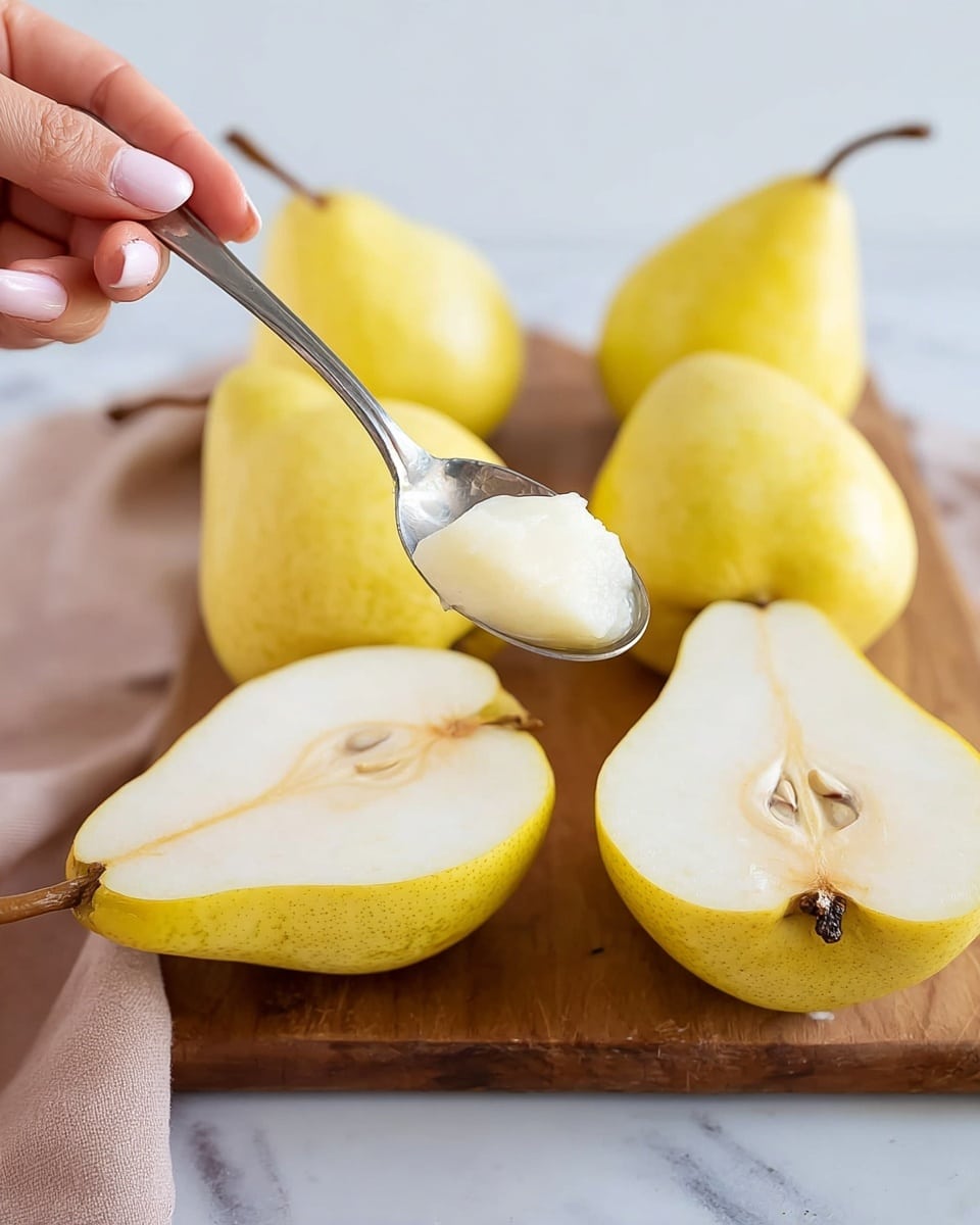 A woman's hand holds a halved yellow pear with soft, light cream flesh. A silver spoon scoops out part of the soft center near the seeds, showing a smooth, creamy texture inside. Around the pear, on a wooden cutting board, are six more pear halves with a similar color and texture, all placed on a white marbled surface. The background is softly blurred to keep focus on the pear and the scooping action. photo taken with an iphone --ar 4:5 --v 7