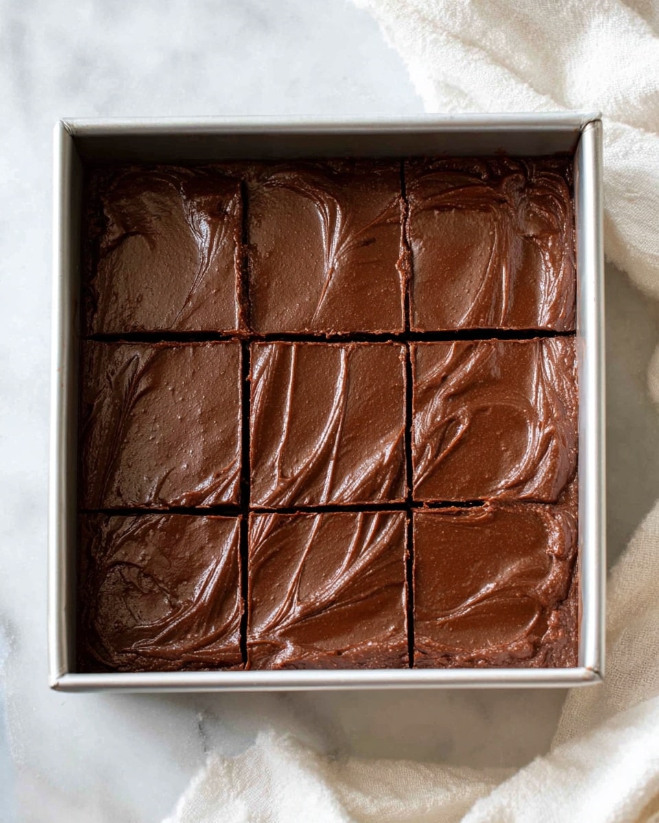 The image shows a square metal baking pan filled with thick, smooth chocolate batter that has been lightly textured with swirls on top. The batter is divided into nine even squares by shallow cut lines, with the rich, dark brown color of the chocolate filling the pan evenly and shining softly in the light. The container rests on a white marbled surface with parts of a white cloth visible in the corner. Photo taken with an iphone --ar 4:5 --v 7