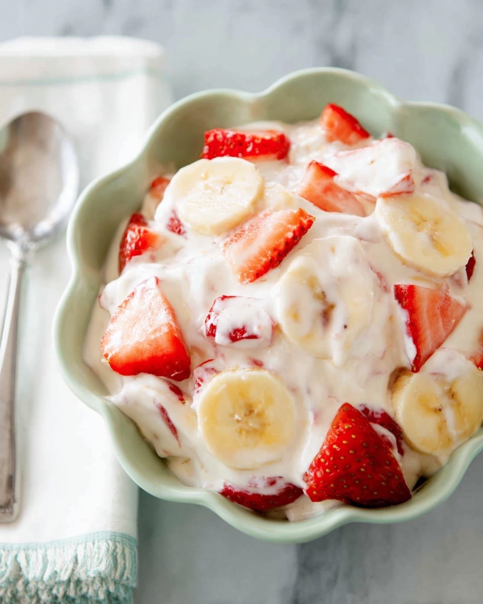 A close-up view of a light green scalloped bowl filled with a creamy white mixture that holds pieces of bright red strawberry slices and pale yellow banana rounds mixed inside. The texture looks soft and smooth with the fruit pieces partially covered by the creamy layer, giving it a fresh and inviting look. The bowl rests on a white marbled surface with part of a white napkin and a metal spoon visible on the side. photo taken with an iphone --ar 4:5 --v 7