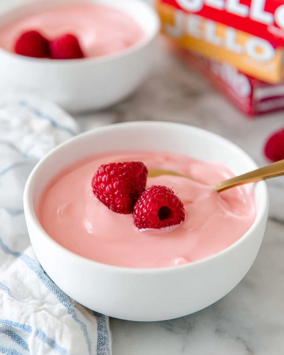 A close-up view of a white bowl filled with soft, smooth pink pudding topped with two fresh red raspberries, with a golden spoon resting inside the bowl on the right side. In the blurred background, there are colorful boxes of Jello gelatin mix in red and cream colors. The bowl is set on a white marbled surface with a part of a white cloth with blue stripes on the left side. Another identical bowl with the same pink pudding and raspberries is partially visible in the background. Photo taken with an iphone --ar 4:5 --v 7