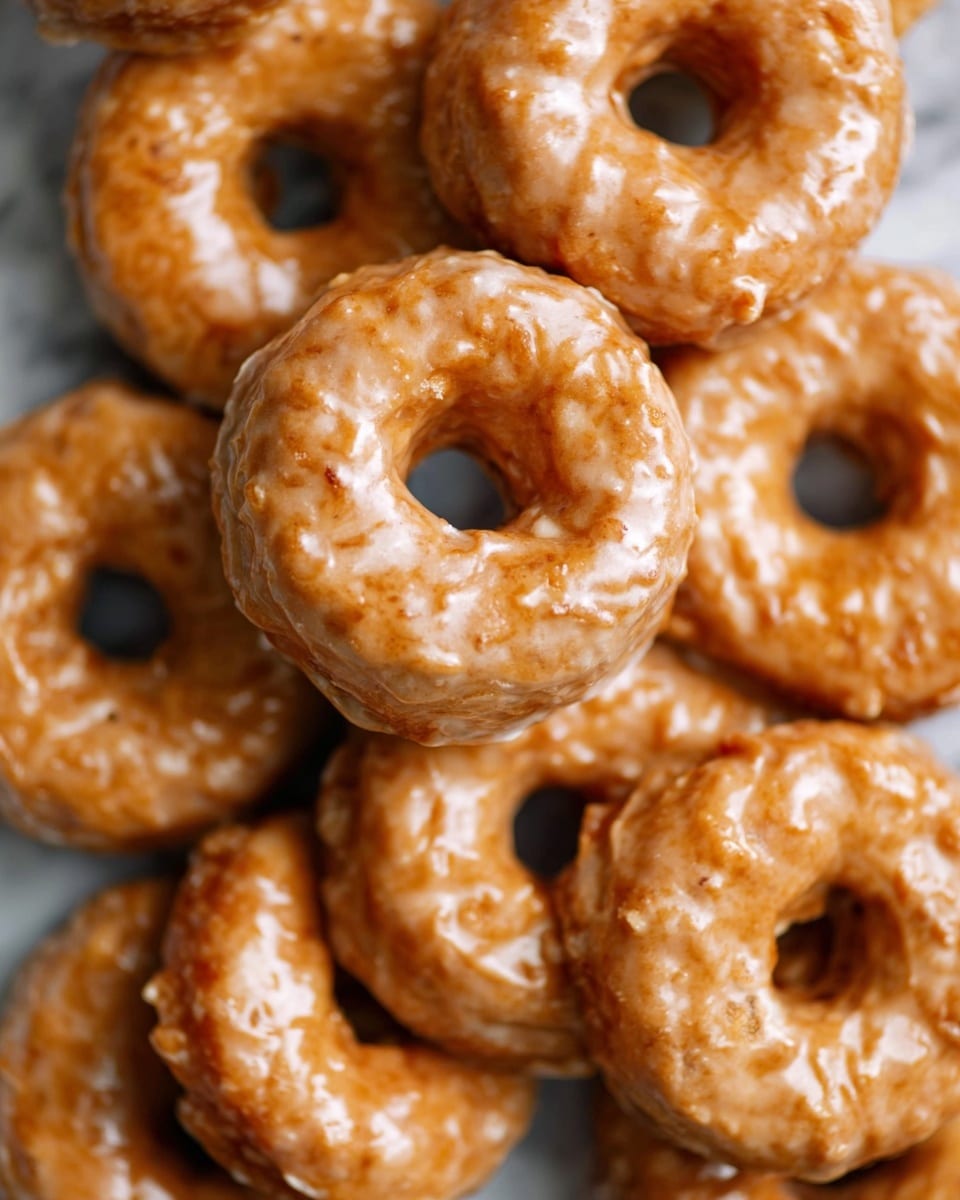 A close-up view of many round donuts with a shiny, light brown glaze covering their rough textured surface. Each donut has a visible hole in the center and they are stacked closely together on a white marbled surface, showing some slight cracks and bumps on the glaze. The lighting highlights the glossy finish, making the donuts look fresh and appetizing. photo taken with an iphone --ar 4:5 --v 7