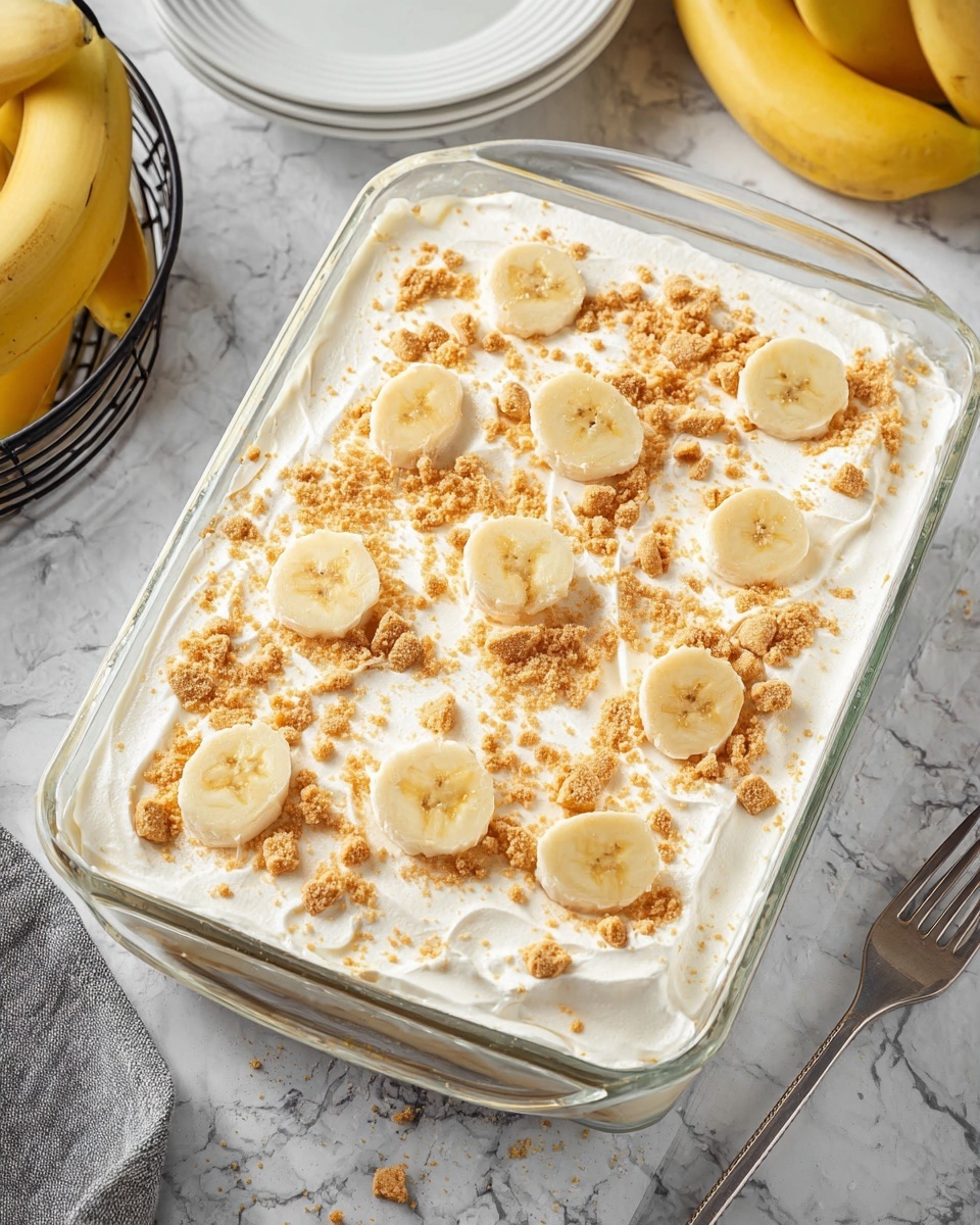 The image shows a clear rectangular glass dish filled with a dessert that has a smooth, white creamy layer on top, likely whipped cream or a similar topping. Scattered over the creamy top are small chunks and crumbs of light brown cookie or biscuit, adding a crunchy texture. Arranged evenly across the surface are several banana slices, each pale yellow with a soft texture. The dish sits on a white marbled surface, with some bananas in a black wire basket visible in the corner. A white plate with a fork is also partially visible nearby. Photo taken with an iphone --ar 4:5 --v 7