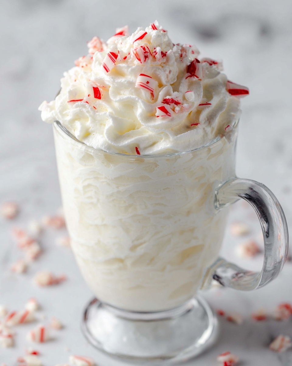 Peppermint Whipped Cream Recipe 4 In a clear glass mug, there is a tall stack of white whipped cream filling the entire glass and overflowing at the top. The whipped cream has a soft, fluffy texture with swirls and peaks. On the top layer, small pieces of red and white striped candy are scattered, adding bright pops of color and a crunchy texture. The mug is placed on a white marbled surface, which adds a clean and elegant background. photo taken with an iphone --ar 4:5 --v 7