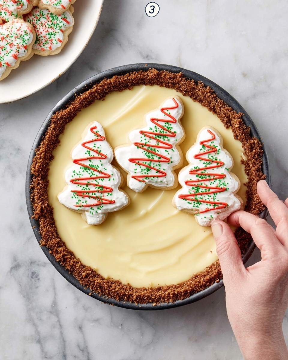 A round pie with a crumbly brown crust forms the bottom and sides, filled with a smooth, pale yellow creamy layer. On top of the pie, there are three white Christmas tree-shaped cookies decorated with red icing zigzags and green sprinkles. A woman's hand is holding one of the cookies, about to place it onto the pie. The pie sits on a white marbled surface with a glimpse of a white plate holding more decorated cookies in the upper left corner. Photo taken with an iphone --ar 4:5 --v 7