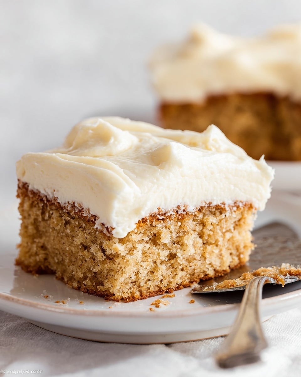 The image shows a close-up of a single square piece of cake with two layers. The bottom layer is thick, moist, and light brown with a slightly crumbly texture. The top layer is a thick, creamy white frosting that looks smooth with soft peaks. The cake rests on a white plate with a slightly raised rim, placed on a white marbled surface. A metal spatula with some crumbs on it is partially visible at the bottom right corner. photo taken with an iphone --ar 4:5 --v 7