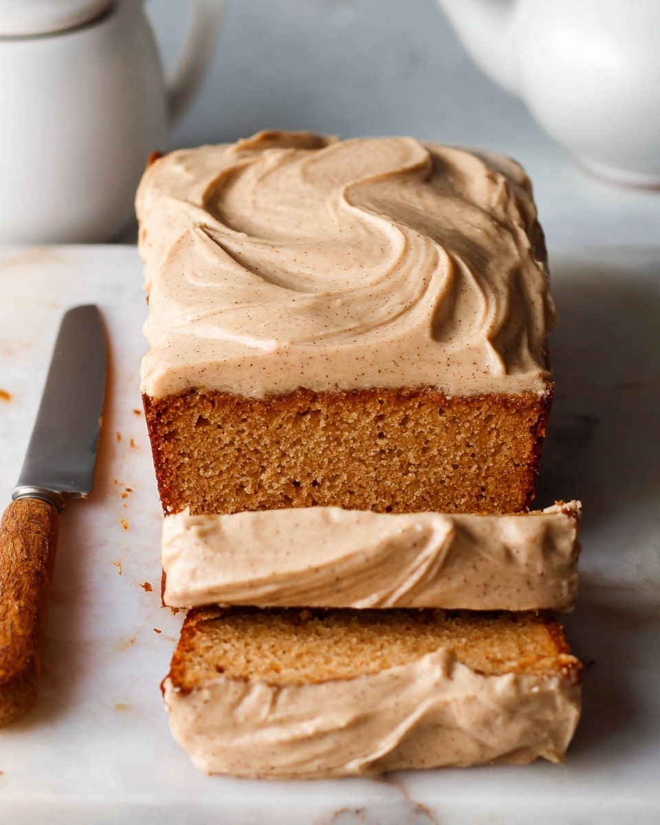 A loaf of cake with thick, creamy light brown frosting swirled smoothly on the top, showing soft, fluffy texture with tiny specks of darker brown throughout. The loaf is sliced into three pieces at the bottom, each piece revealing a moist, golden brown cake layer underneath the frosting. The cake sits on a white marbled surface, with a silver knife with a wooden handle placed beside it. A white teapot is visible in the background at the top edge of the image. Photo taken with an iphone --ar 4:5 --v 7