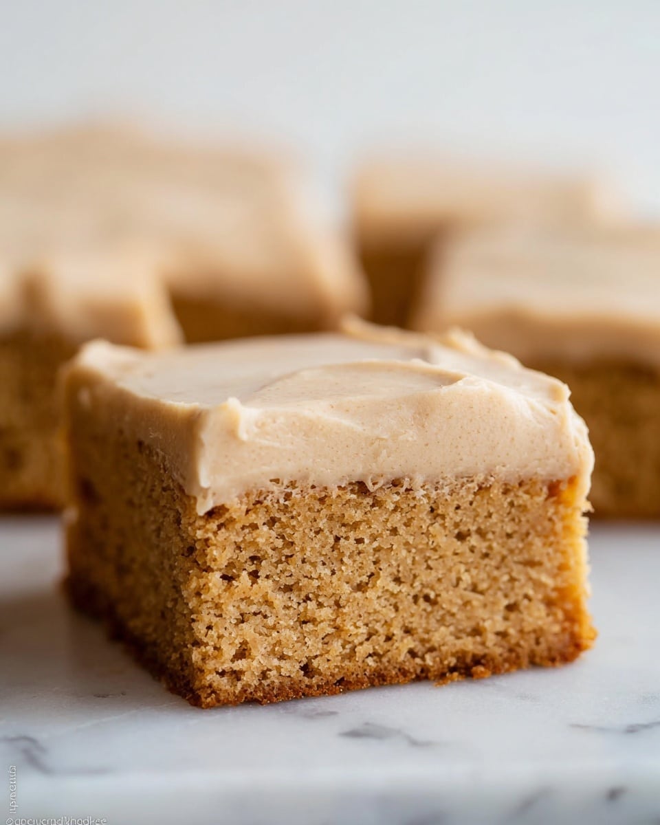 The image shows a close-up of a square piece of two-layer cake on a white marbled surface. The bottom layer is a light brown, moist, and crumbly cake with a slightly rough texture made visible by tiny air holes, while the top layer is a thick, creamy frosting of beige color with a smooth but slightly uneven surface. The edges of the cake are cleanly cut, and in the background, more similar pieces are visible, slightly blurred. photo taken with an iphone --ar 4:5 --v 7