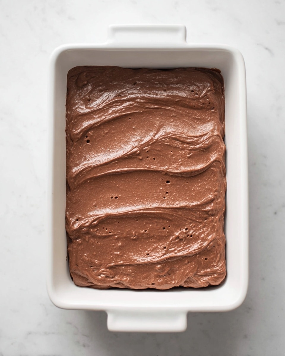 A white rectangular baking dish filled with one thick layer of smooth, creamy chocolate batter spread evenly across the dish, showing soft ridges and small air bubbles on the surface. The dish is placed on a white marbled texture background. photo taken with an iphone --ar 4:5 --v 7
