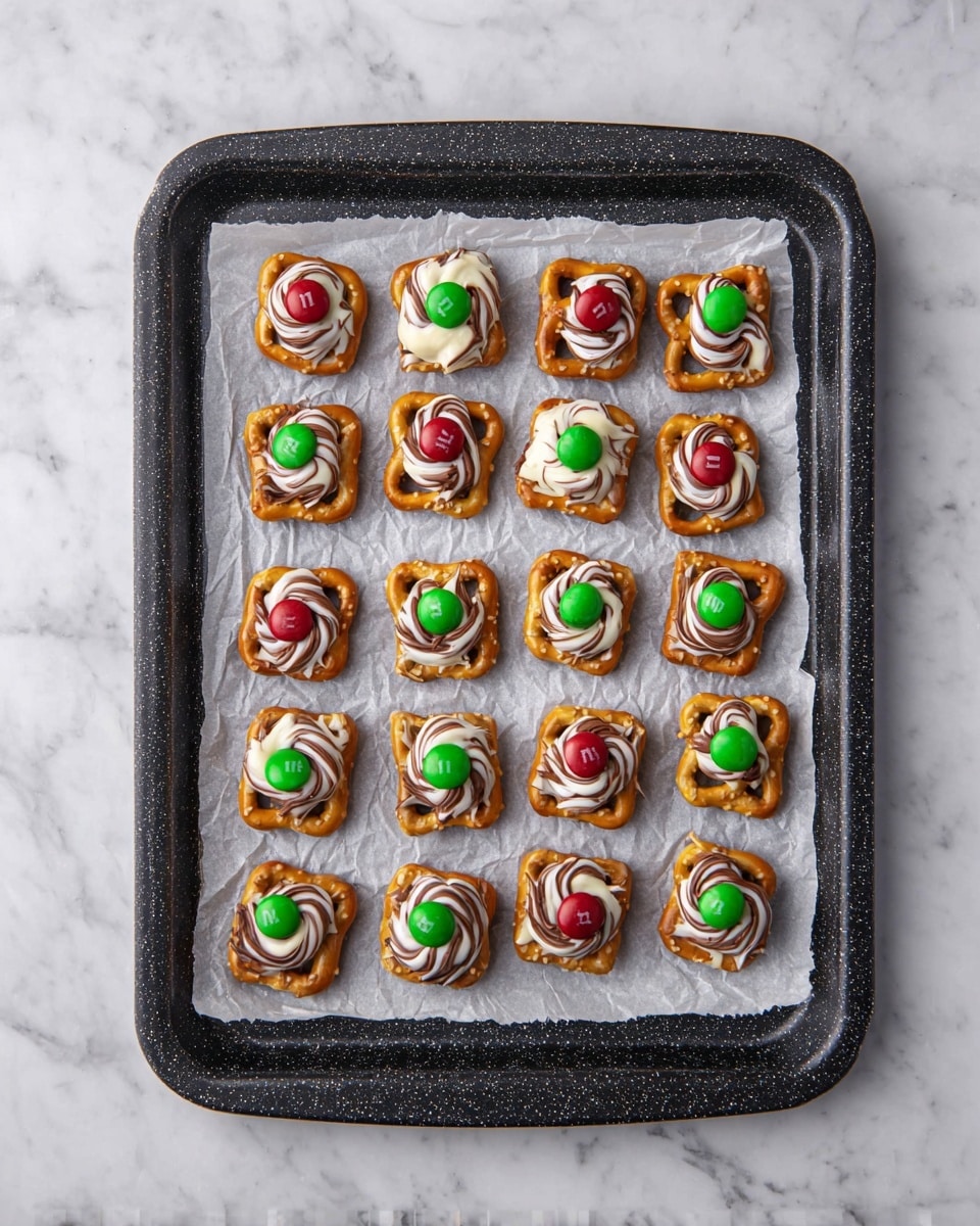 The image shows a black speckled baking tray lined with a crumpled white parchment paper, resting on a white marbled surface. On the tray, 25 small square pretzels are arranged in a 5 by 5 grid. Each pretzel has one of two types of toppings: half of them are topped with a swirl of white and brown chocolate with a green M&M candy in the center, while the other half have a dollop of milk chocolate with either a red or green M&M candy placed neatly on top. The layers are clearly visible with the twisted lines of chocolate contrasting against the square pretzels and shiny smooth candy centered on each piece. photo taken with an iphone --ar 4:5 --v 7