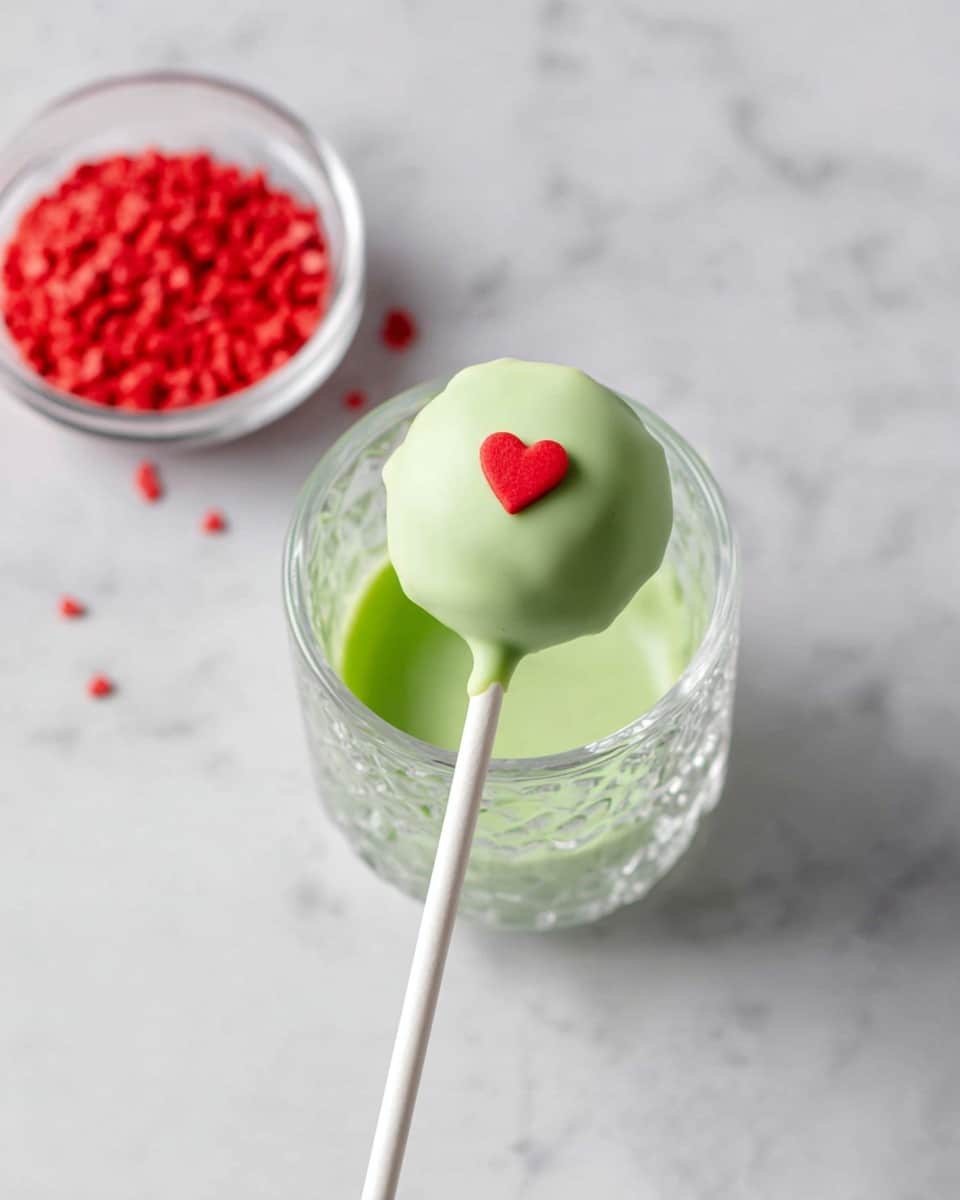 A round cake pop covered in smooth light green coating sits on a white stick. It has a small red heart-shaped decoration placed on top in the middle. The cake pop is dipped in a textured clear glass filled with the same light green coating. In the upper left corner, a clear bowl holds red heart-shaped sprinkles. All of this is on a white marbled surface. photo taken with an iphone --ar 4:5 --v 7